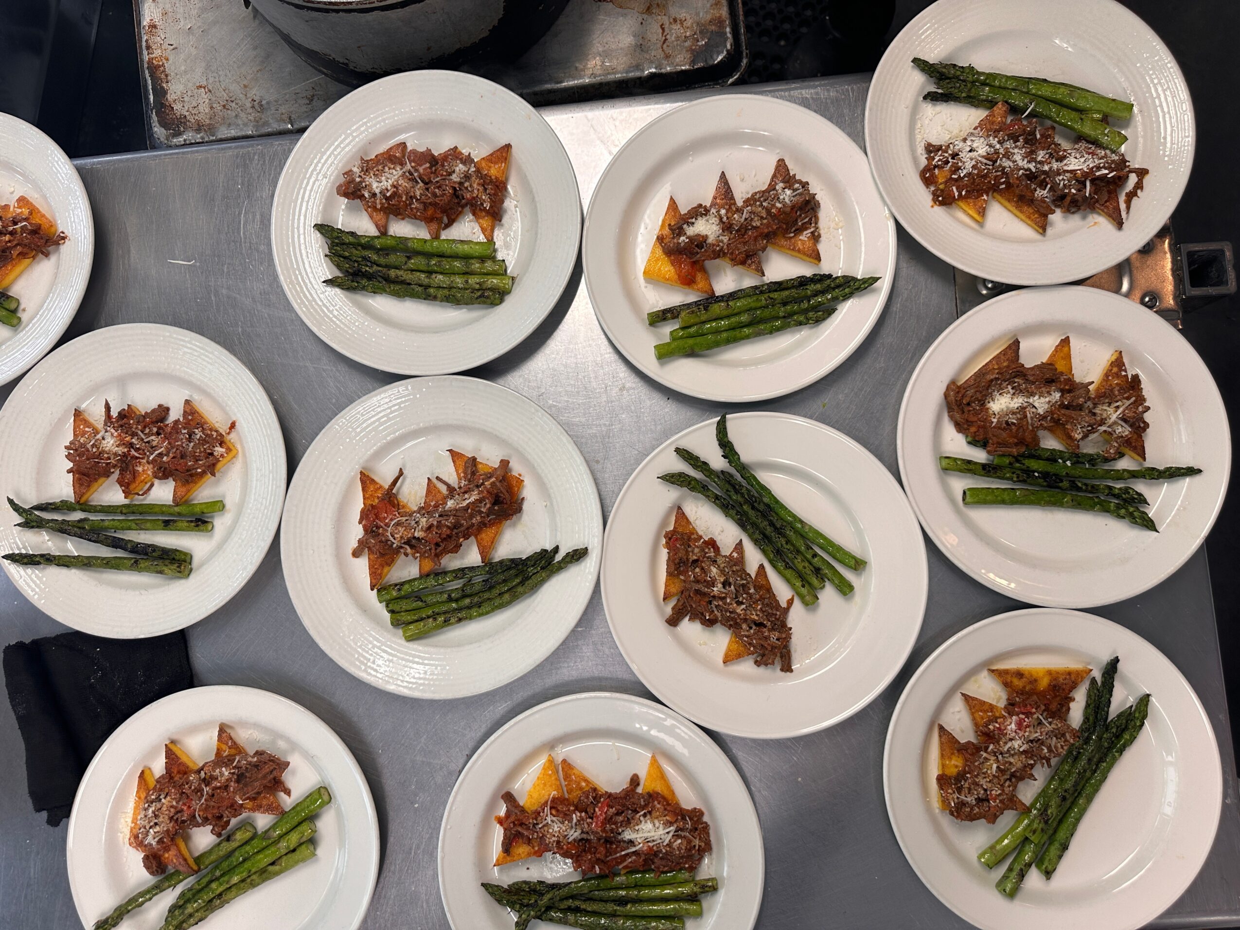 Multiple white plates with fish, green beans, and lemon wedges on a stainless steel surface.