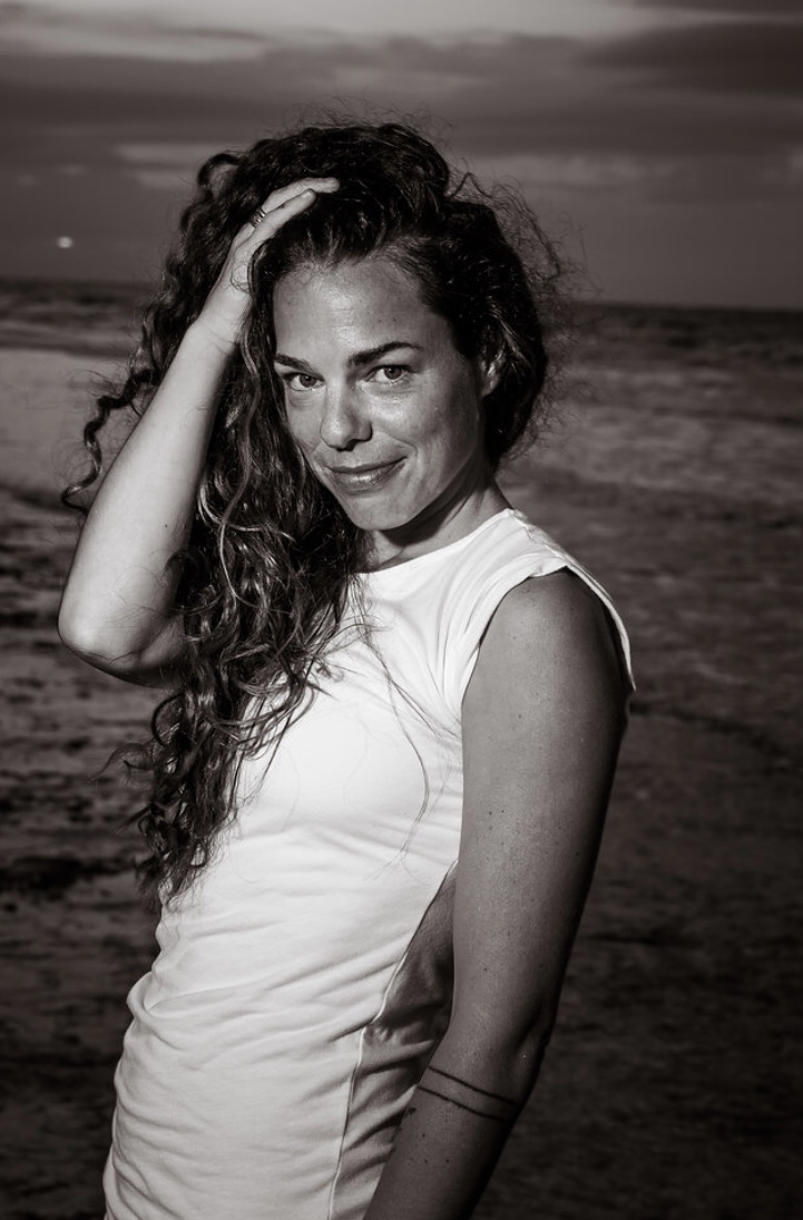 Woman with long curly hair on a beach, looking at the camera, hand on her head, smiling, sunset in background.