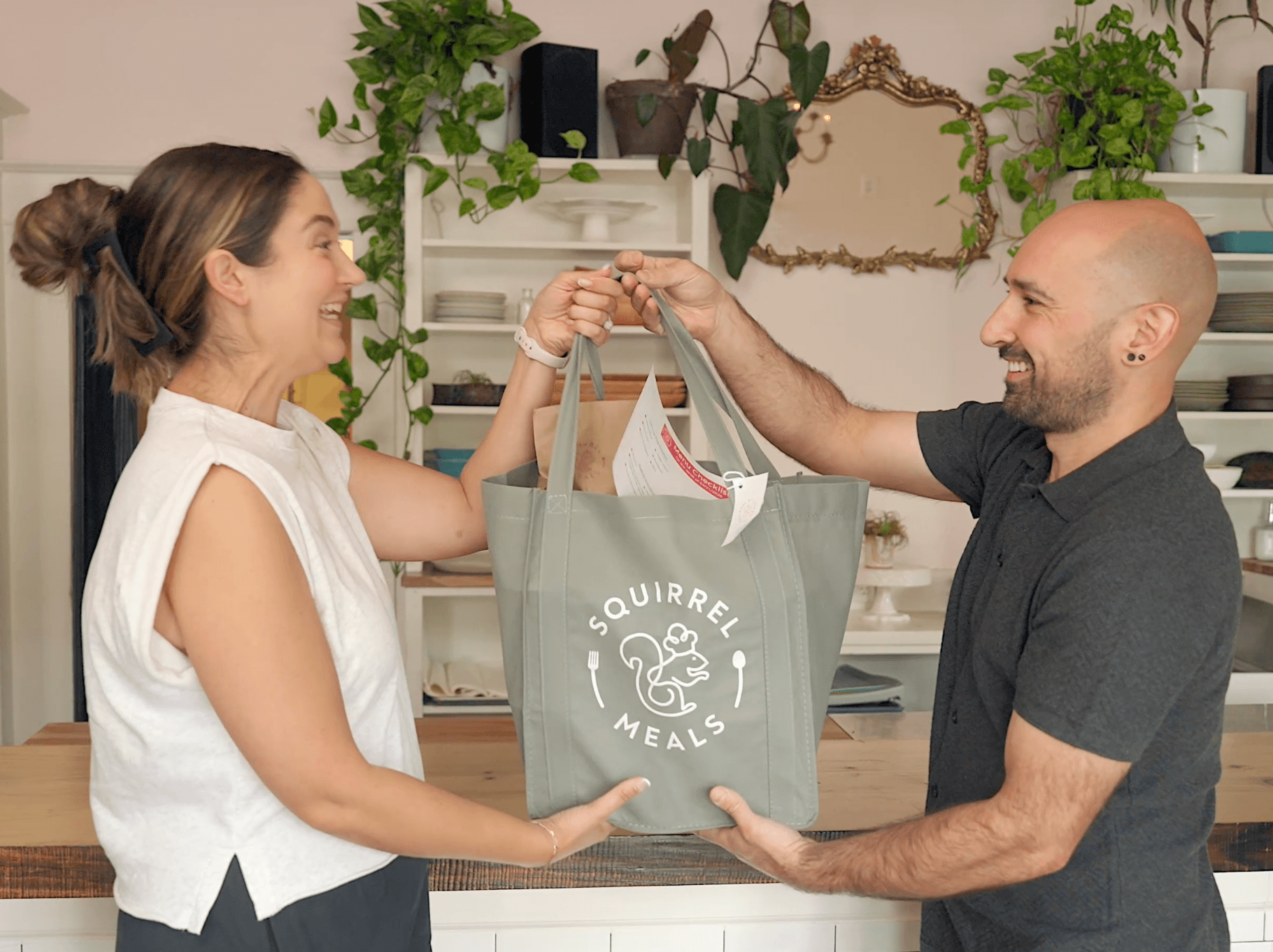 A man hands a woman a reusable shopping bag with a squirrel logo, in a room with plants and shelves.