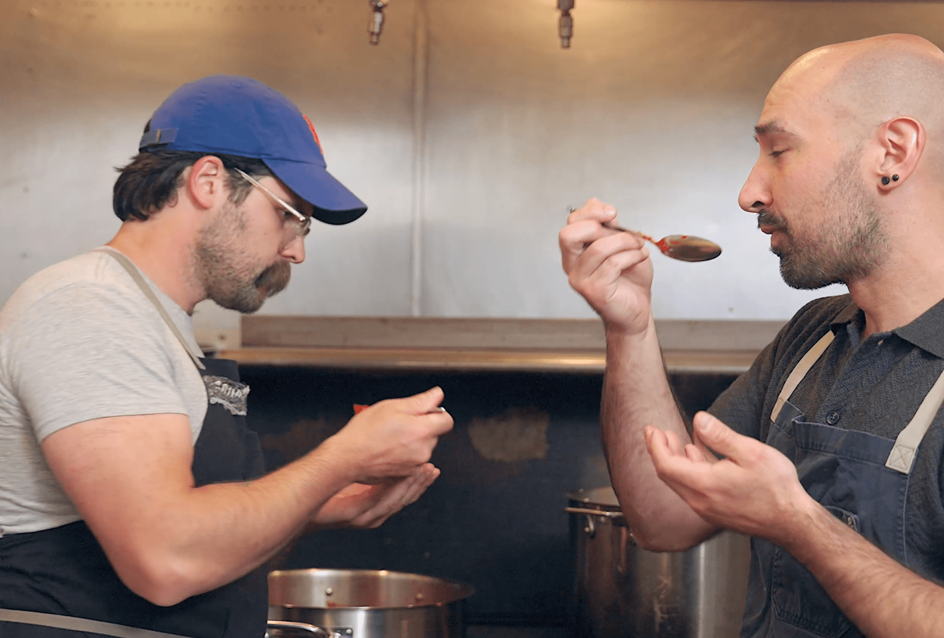 Two men in a kitchen, one wearing a blue cap and glasses, the other bald, tasting food with a spoon.