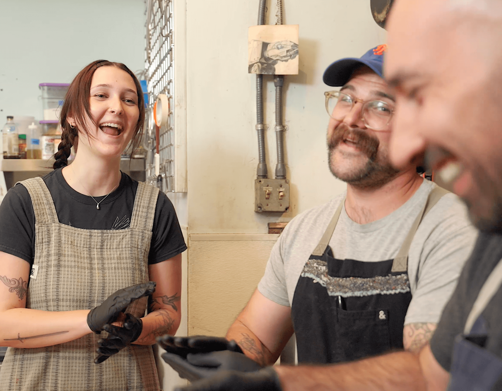 Three people smiling and talking indoors, one woman wearing an apron and gloves, two men with beards, casual clothing.