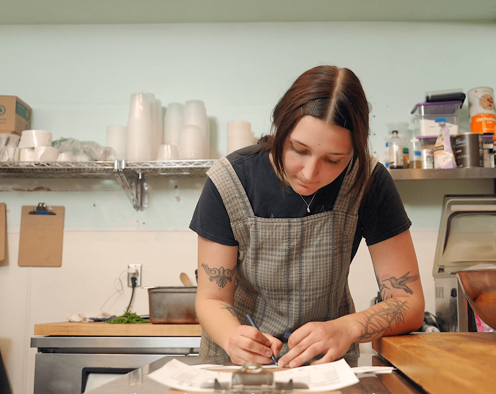 Woman with tattoos writing at a counter in a kitchen or cafe, shelves with cups and supplies behind her.