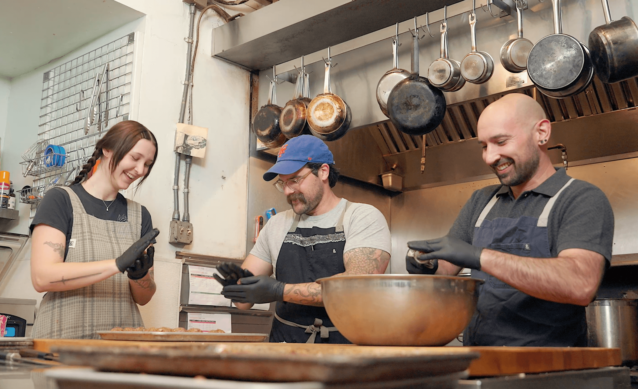 Three people in a kitchen, smiling and looking at tablets, with pots hanging above them.