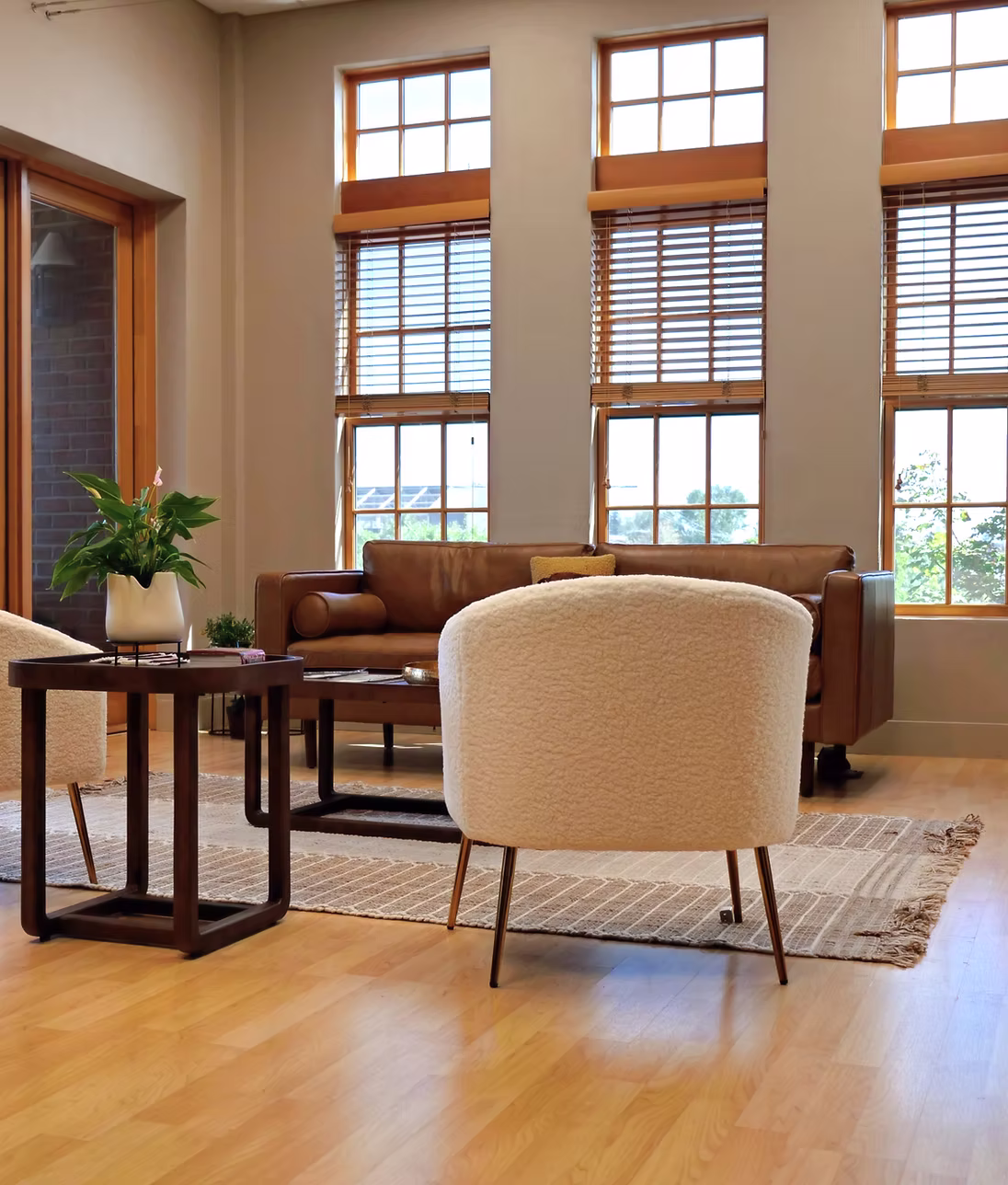 Living room with large windows, brown sofa, beige armchair, wooden side table, potted plant, and a rug on wooden floor.