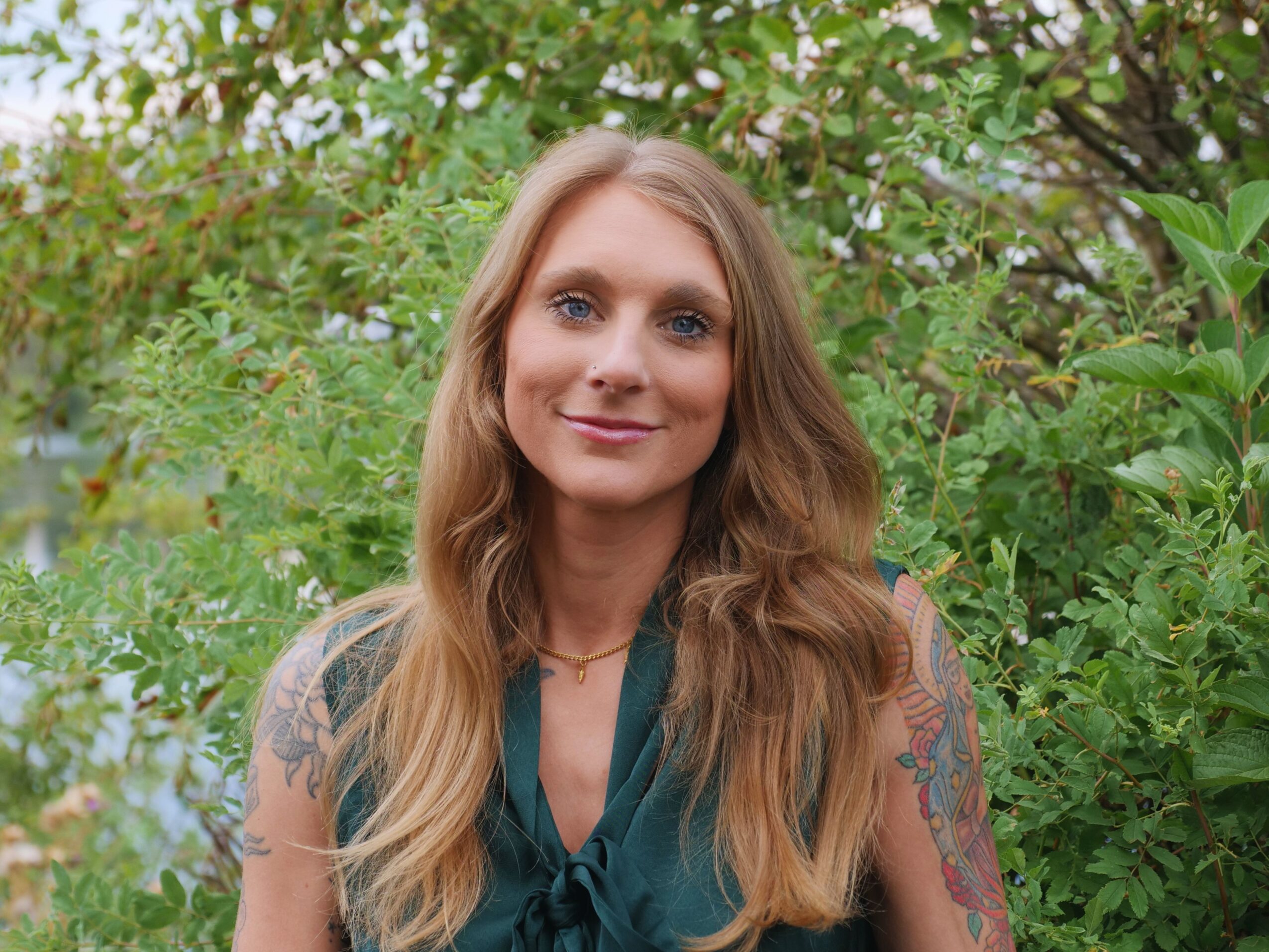 Young woman with long wavy hair smiling outdoors surrounded by green foliage.