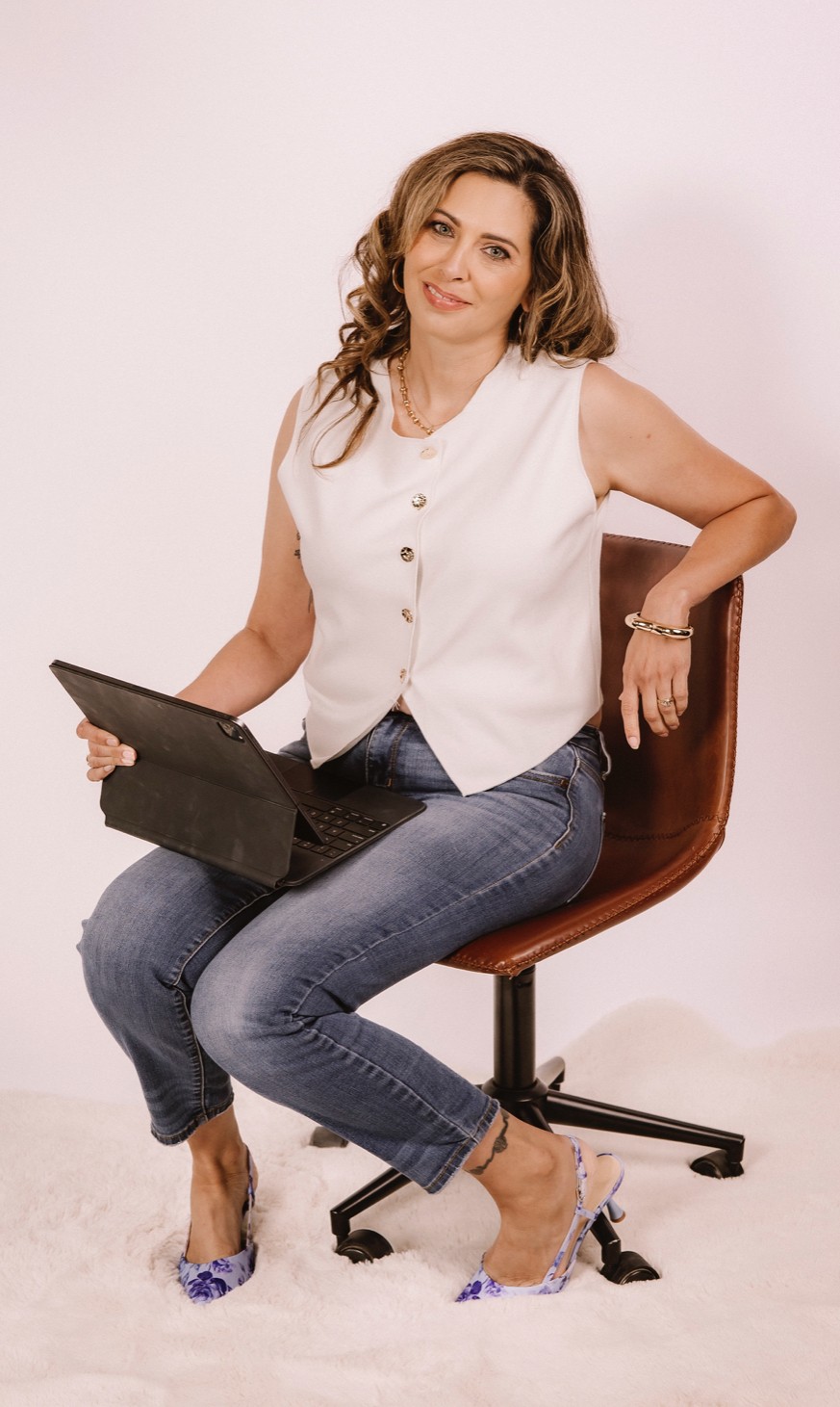 Woman sitting on a chair holding a tablet, smiling, wearing a white sleeveless top and jeans, against a plain background.