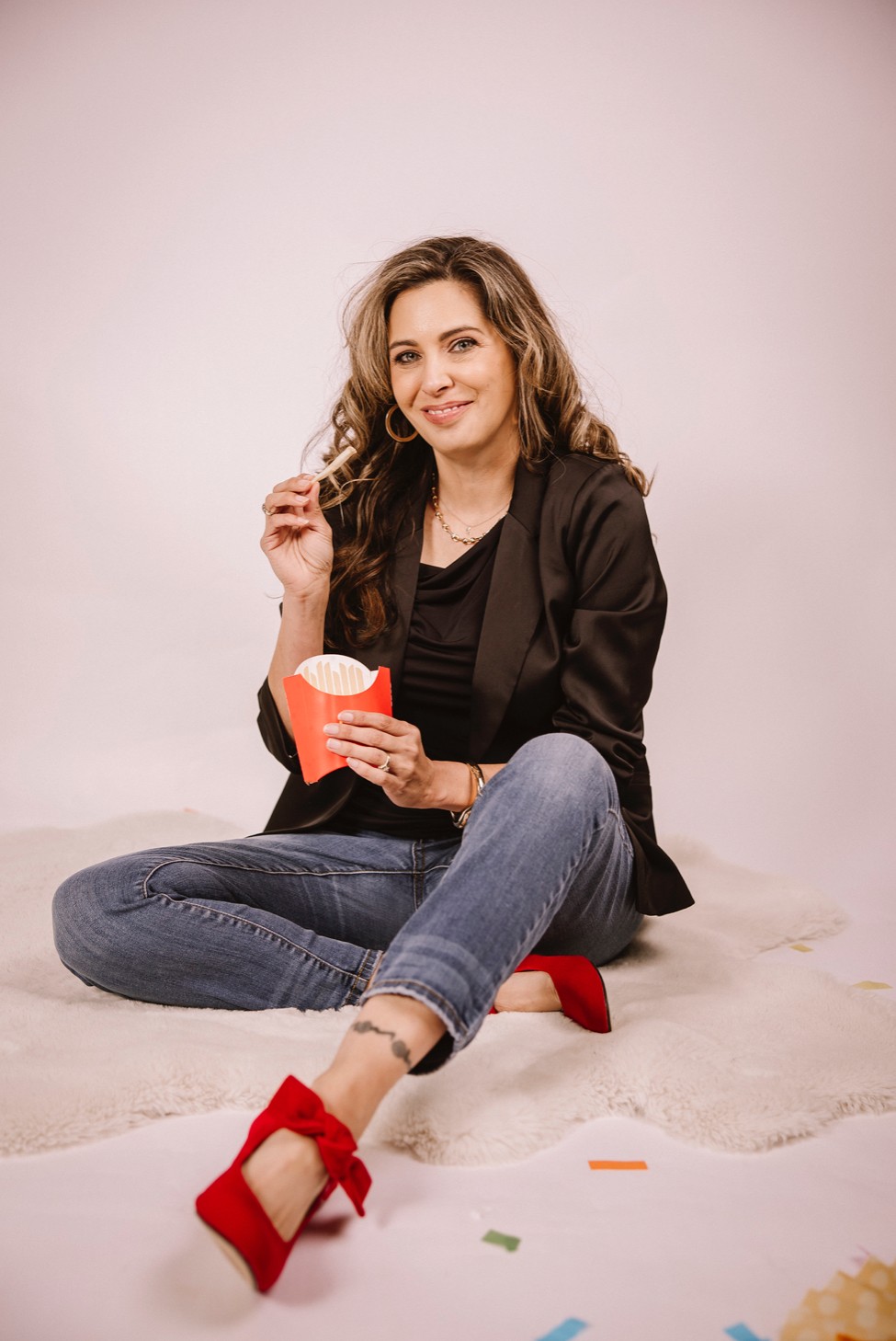 Woman sitting on floor with a popcorn bucket, smiling, wearing a black jacket, jeans, and red shoes, against a plain background.