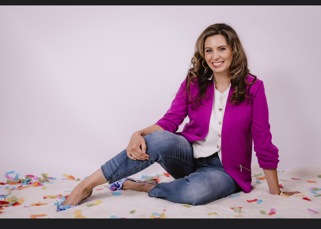 Woman sitting on floor with colorful confetti, smiling, wearing a purple jacket and jeans, against a plain white background.