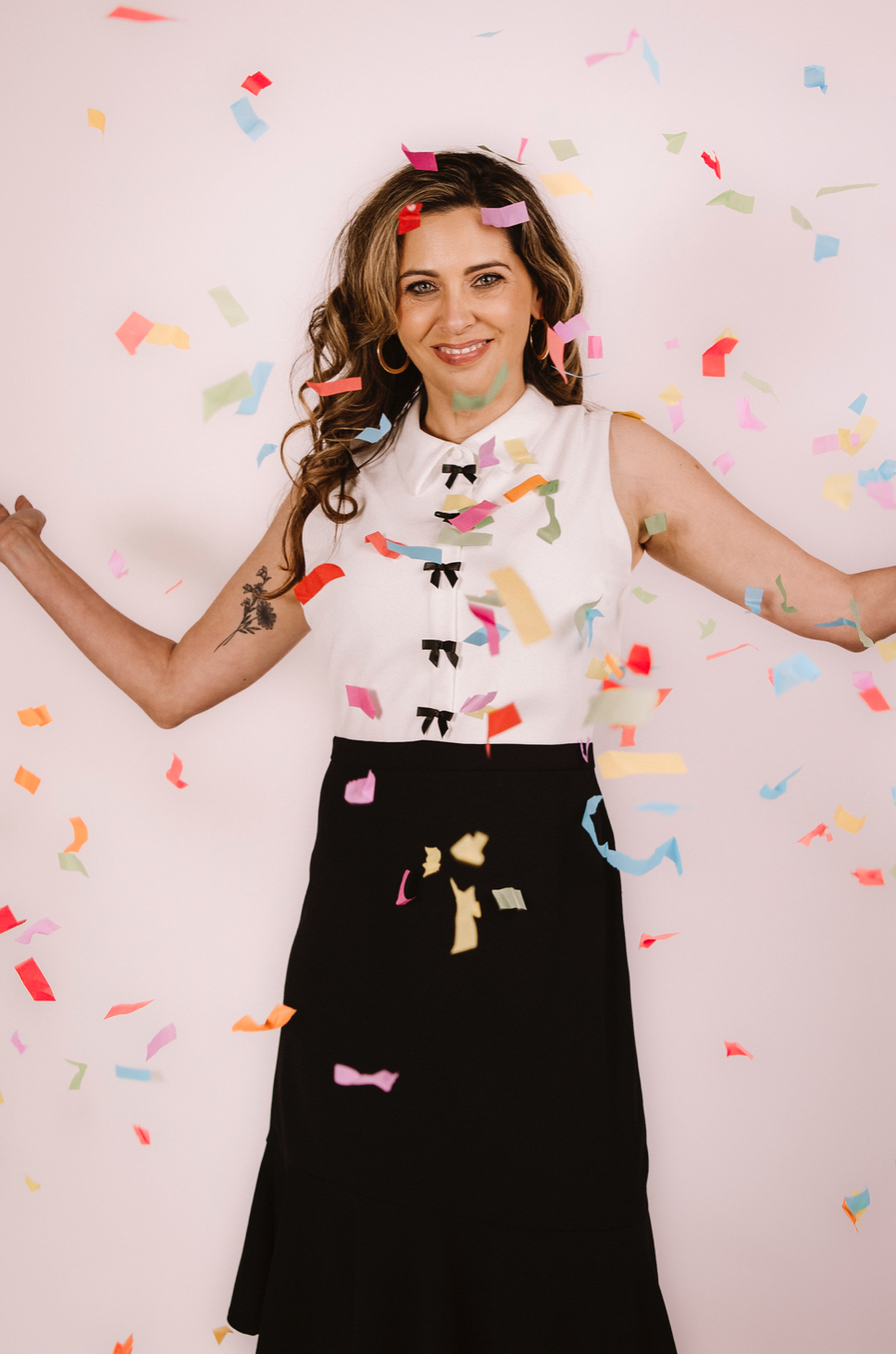 Young woman celebrating with colorful confetti falling around her, smiling, arms outstretched, against a white background.