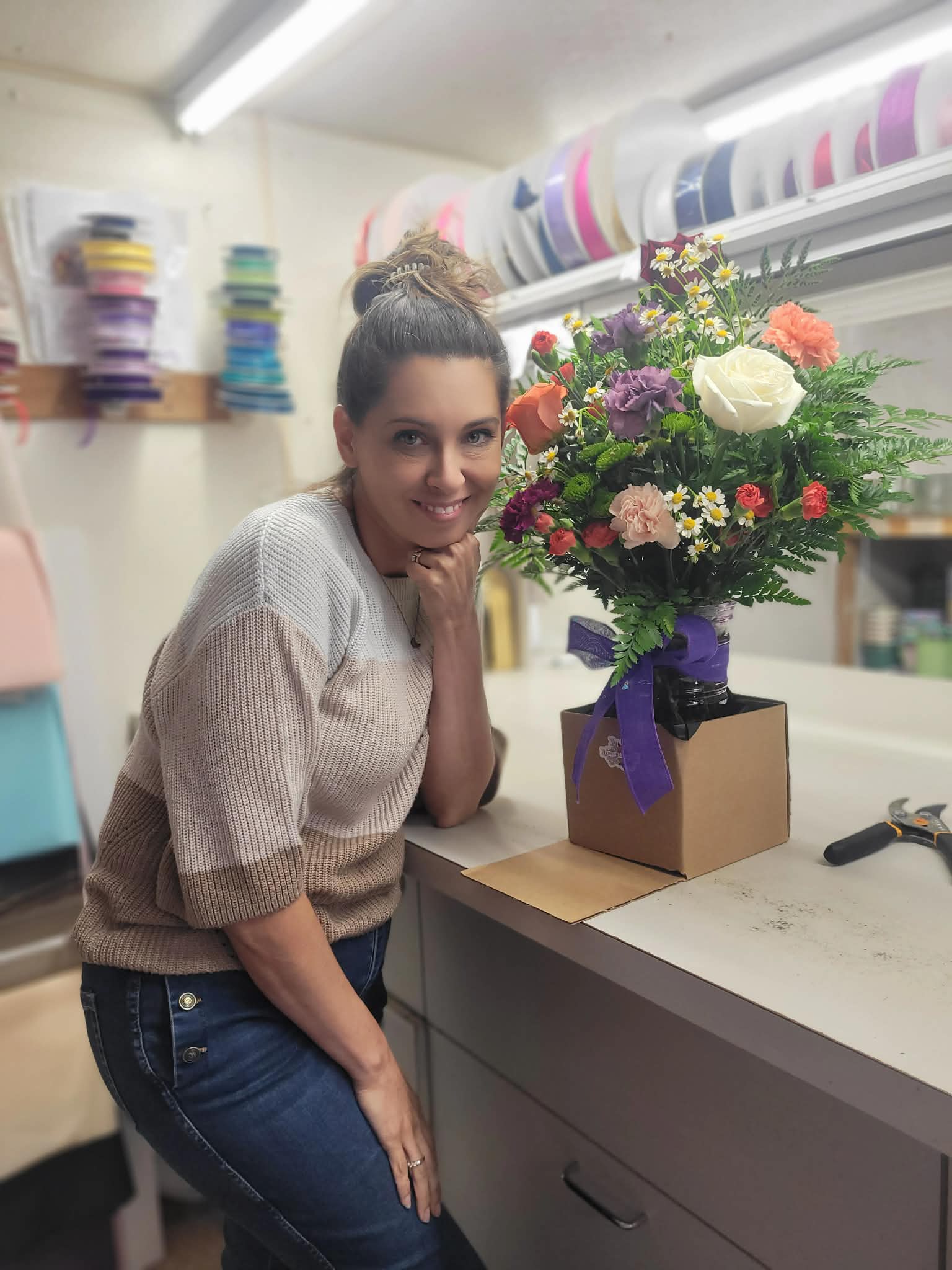 Woman with a bun hairstyle smiling next to a colorful flower bouquet on a counter in a shop or workshop.