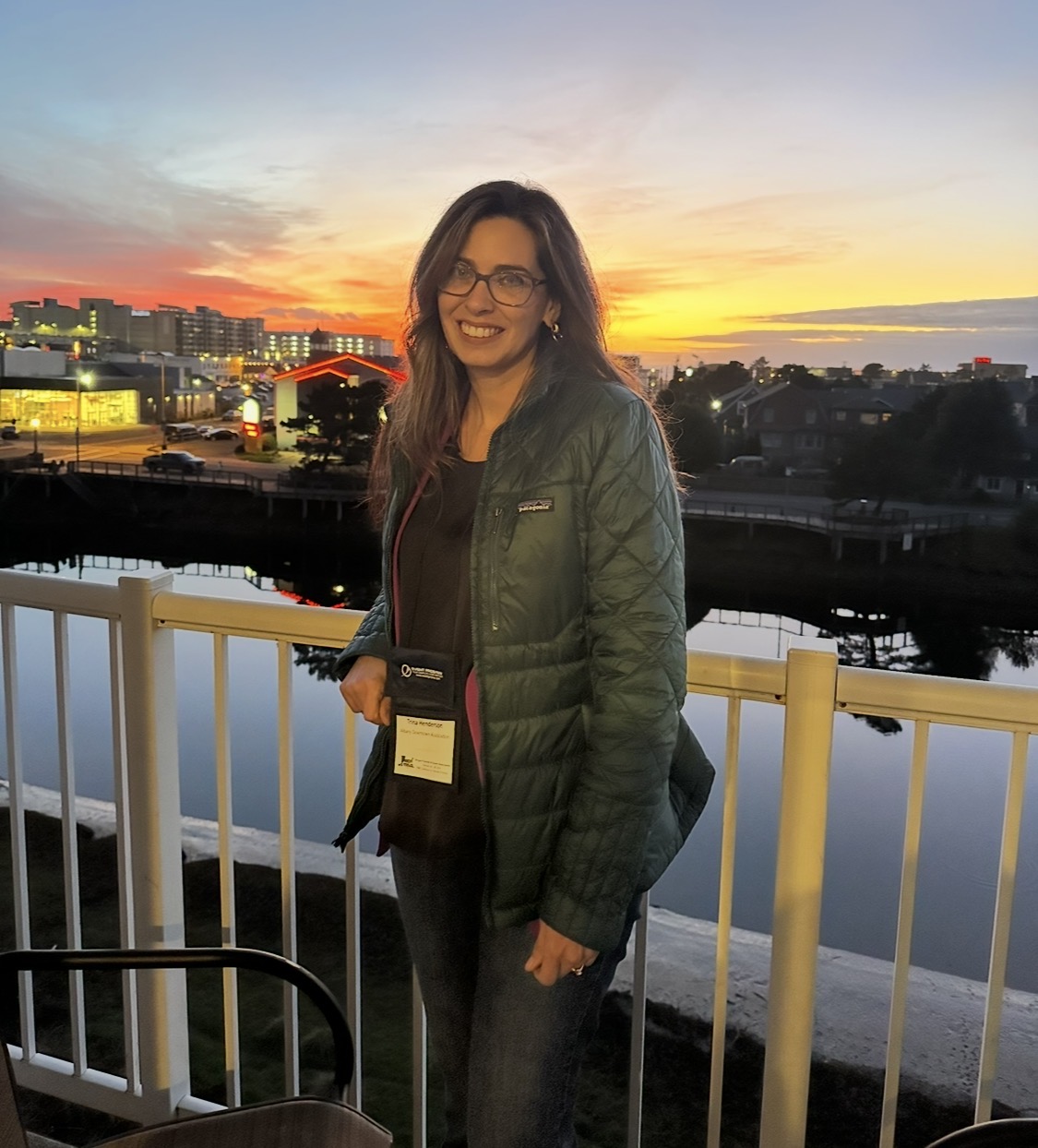 Woman with glasses and long hair standing outdoors near a railing during sunset, smiling, with city buildings and water in background.