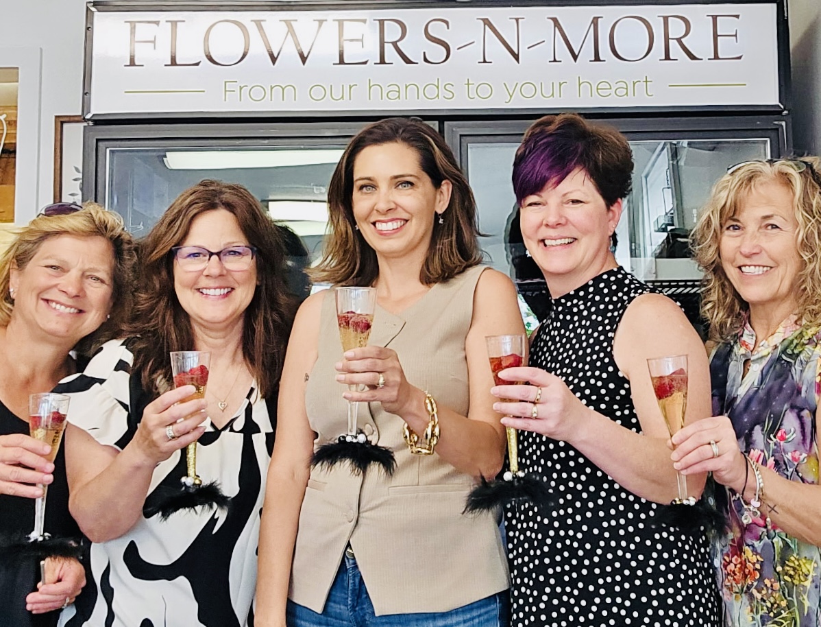 Group of five women smiling and holding drinks in front of a flower shop sign.