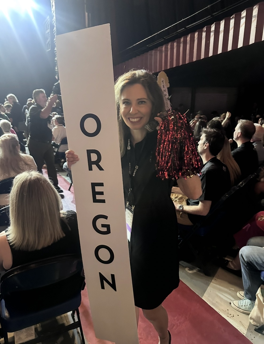 Woman smiling holding a vertical sign with the word 'OREGON' at an indoor event, surrounded by seated and standing people.