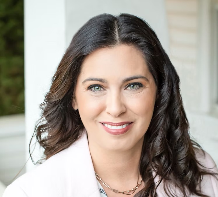 Portrait of a woman with dark wavy hair, smiling, wearing a white blazer, outdoors near a building, natural light.
