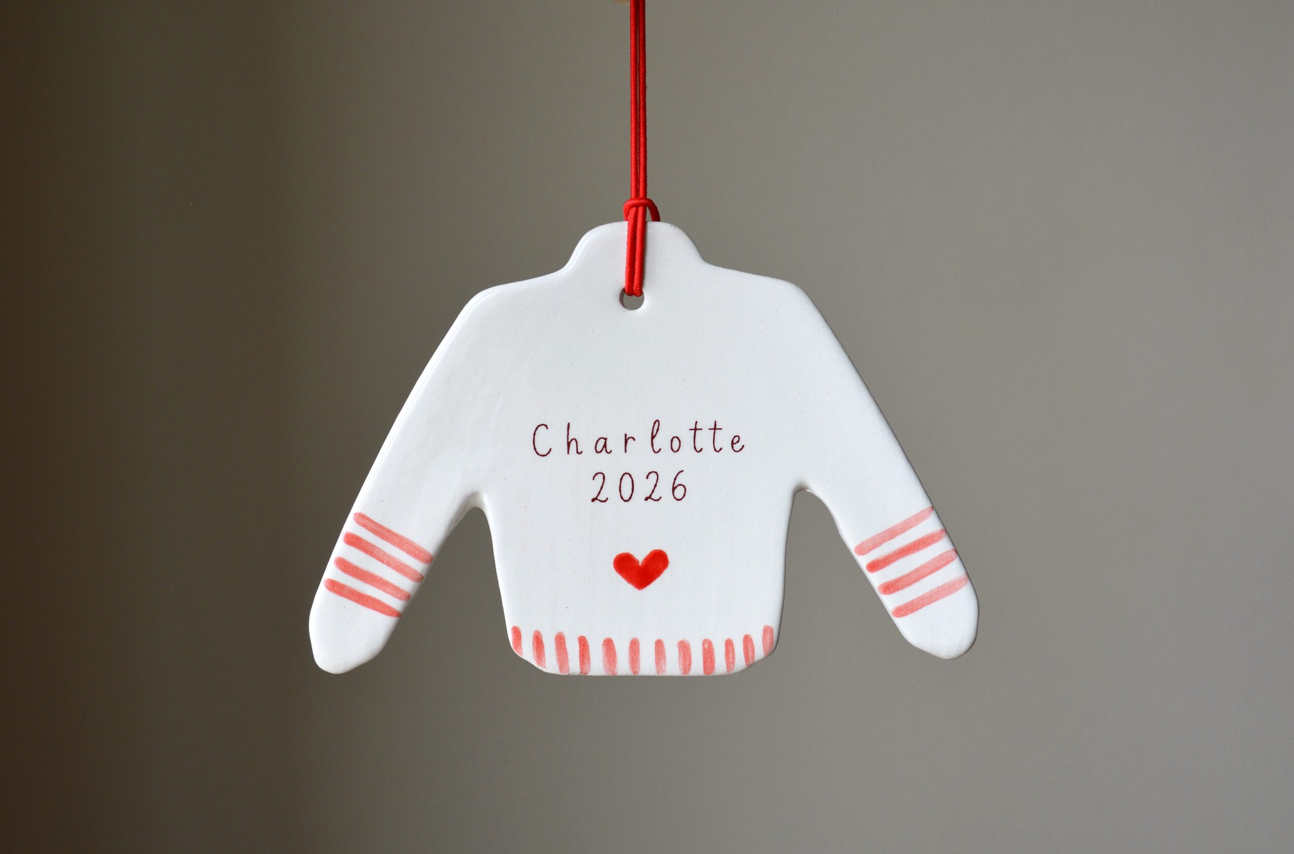 White sweater-shaped ornament with red stripes on sleeves, red heart, and handwritten text, hanging on a red string against a gray background.
