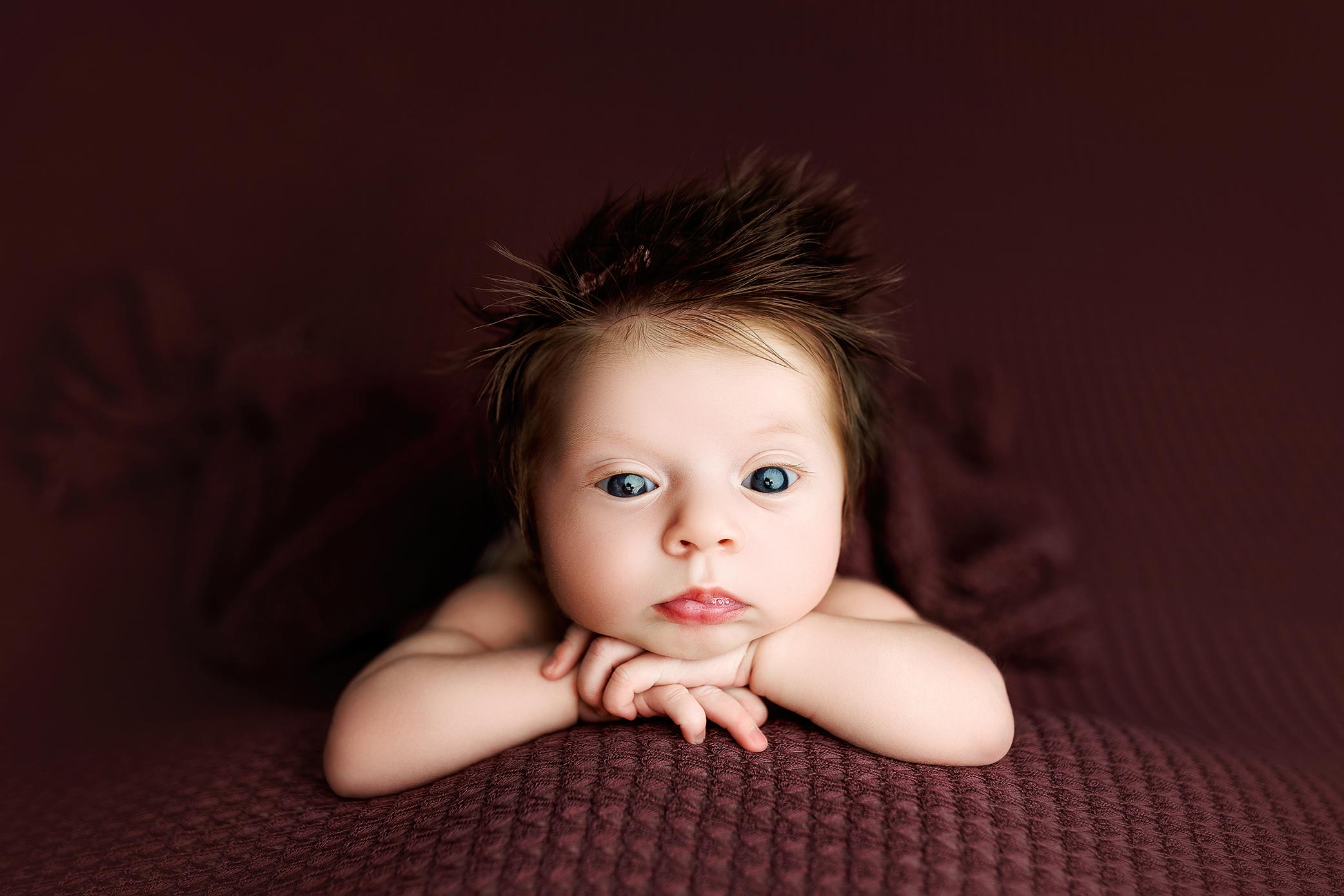 Baby with spiky hair resting chin on hands on dark textured background