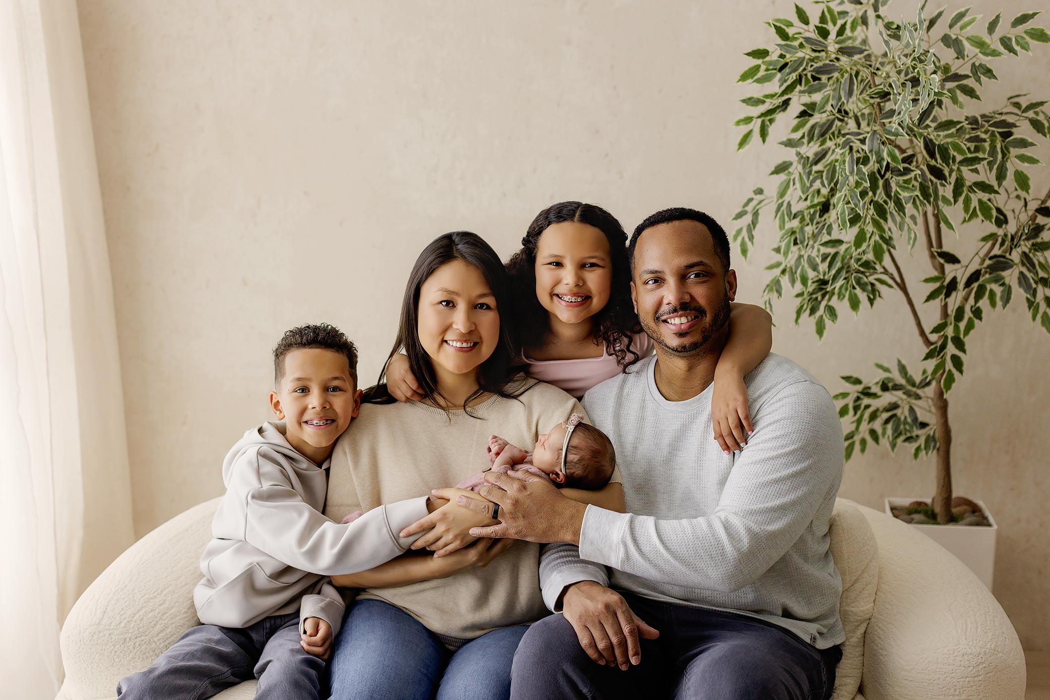 Family of five sitting on a sofa, smiling, with a large plant in the background.