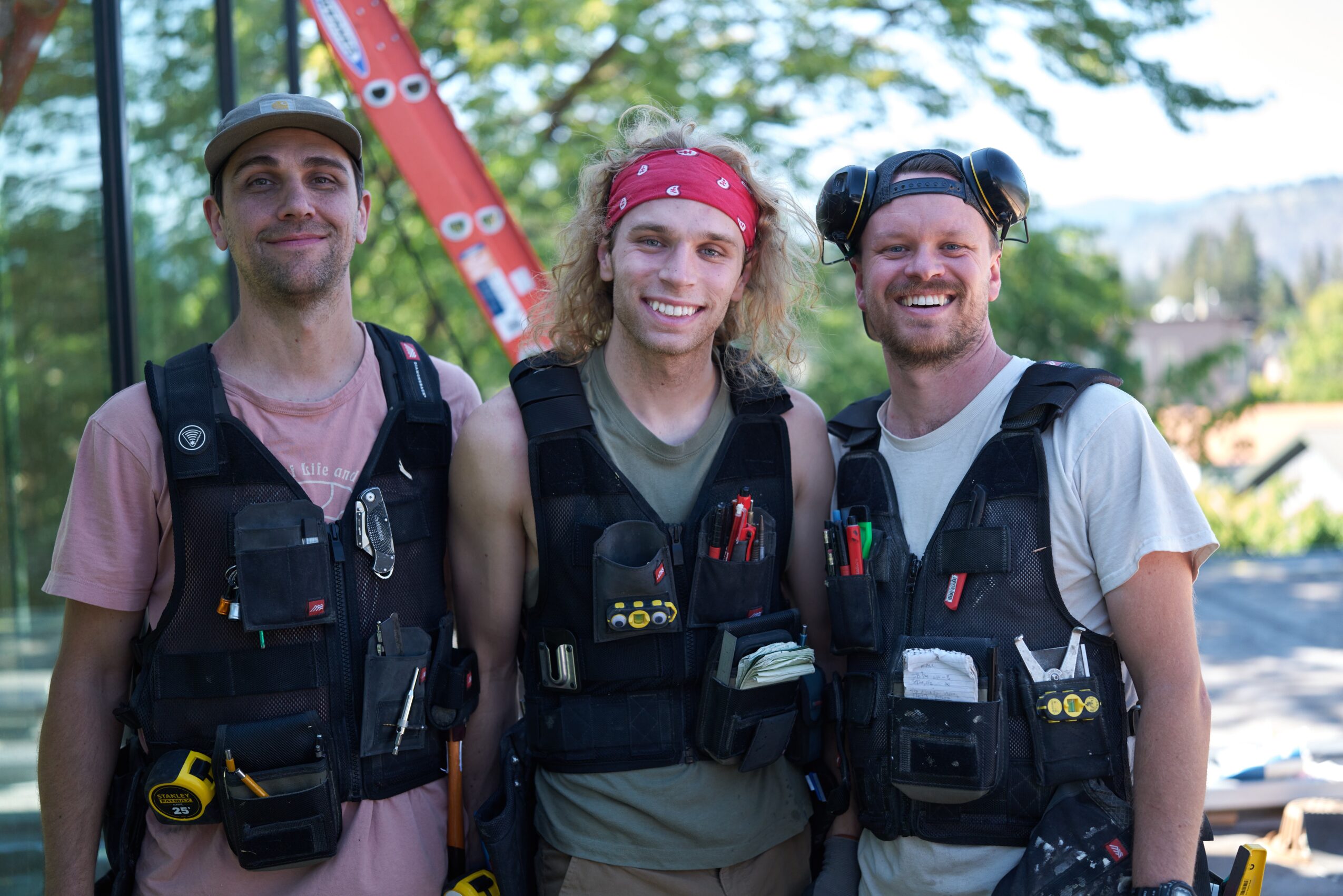 Three people wearing life jackets outdoors, smiling, with trees and a body of water in the background.