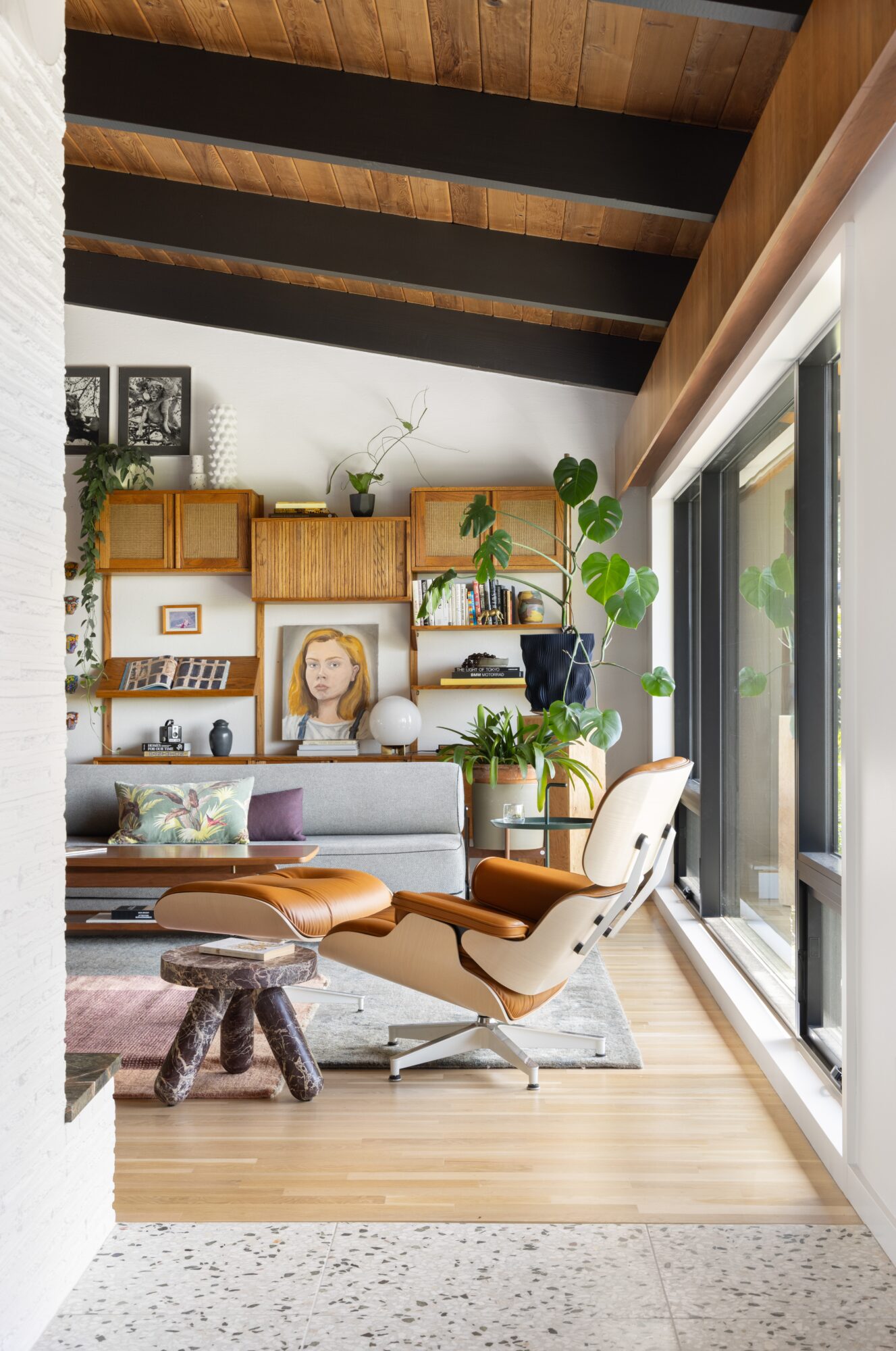 Living room with wooden ceiling beams, bookshelf, plants, sofa, and a white chair near sliding glass doors.