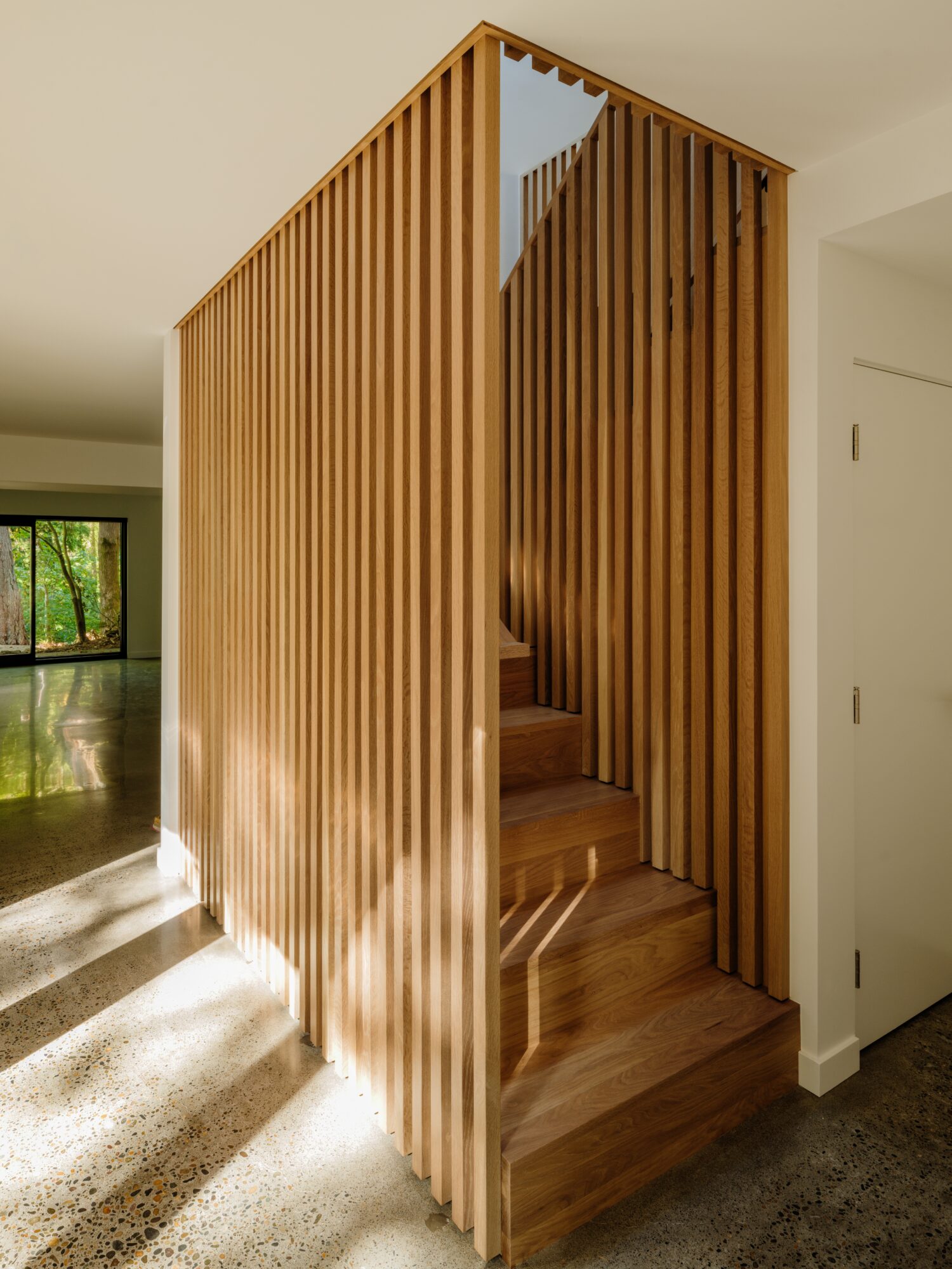 Wooden staircase enclosed by vertical slats, leading upward, with sunlight casting shadows on the floor.
