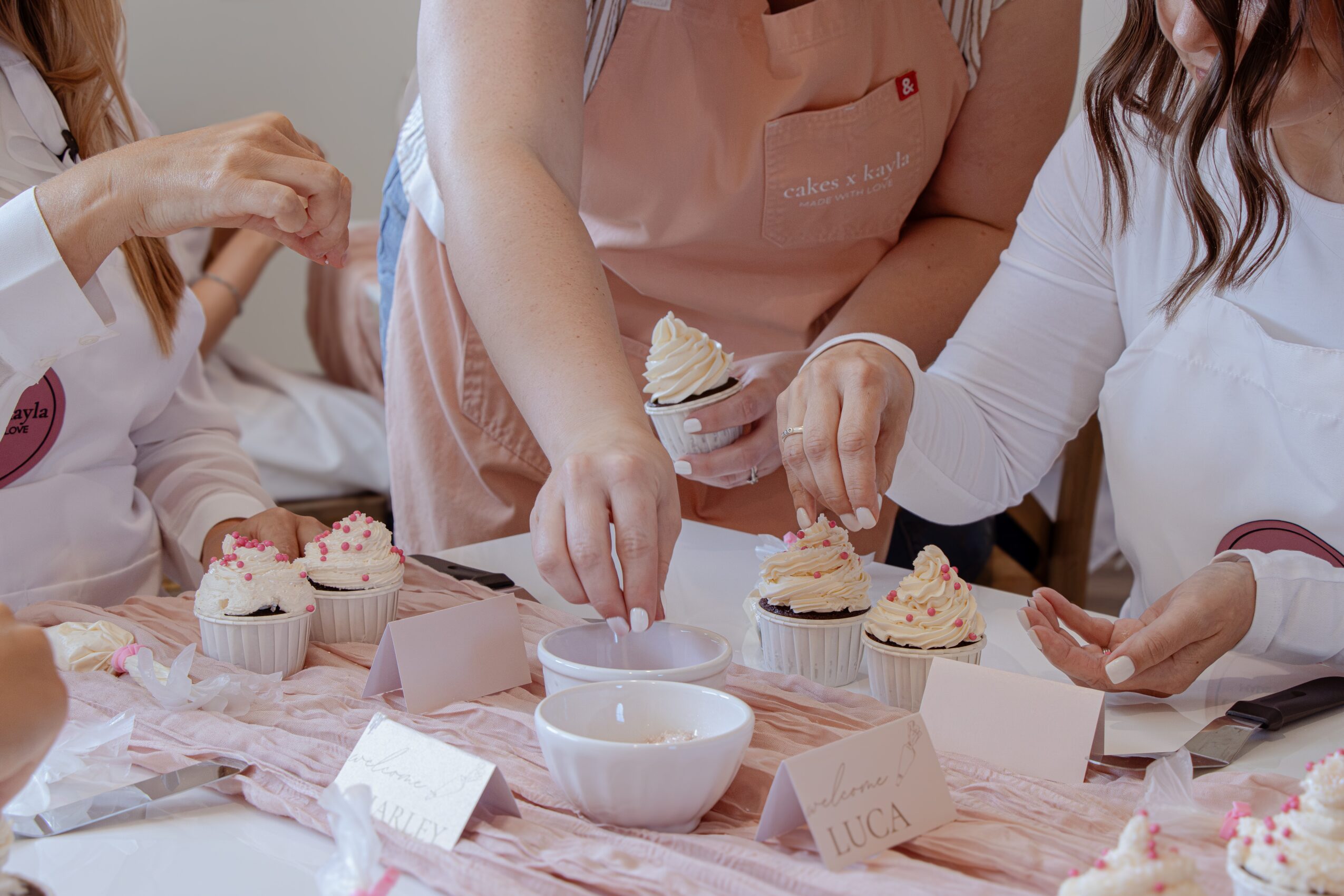 People decorating cupcakes with frosting and sprinkles on a table, with bowls and boxes nearby.