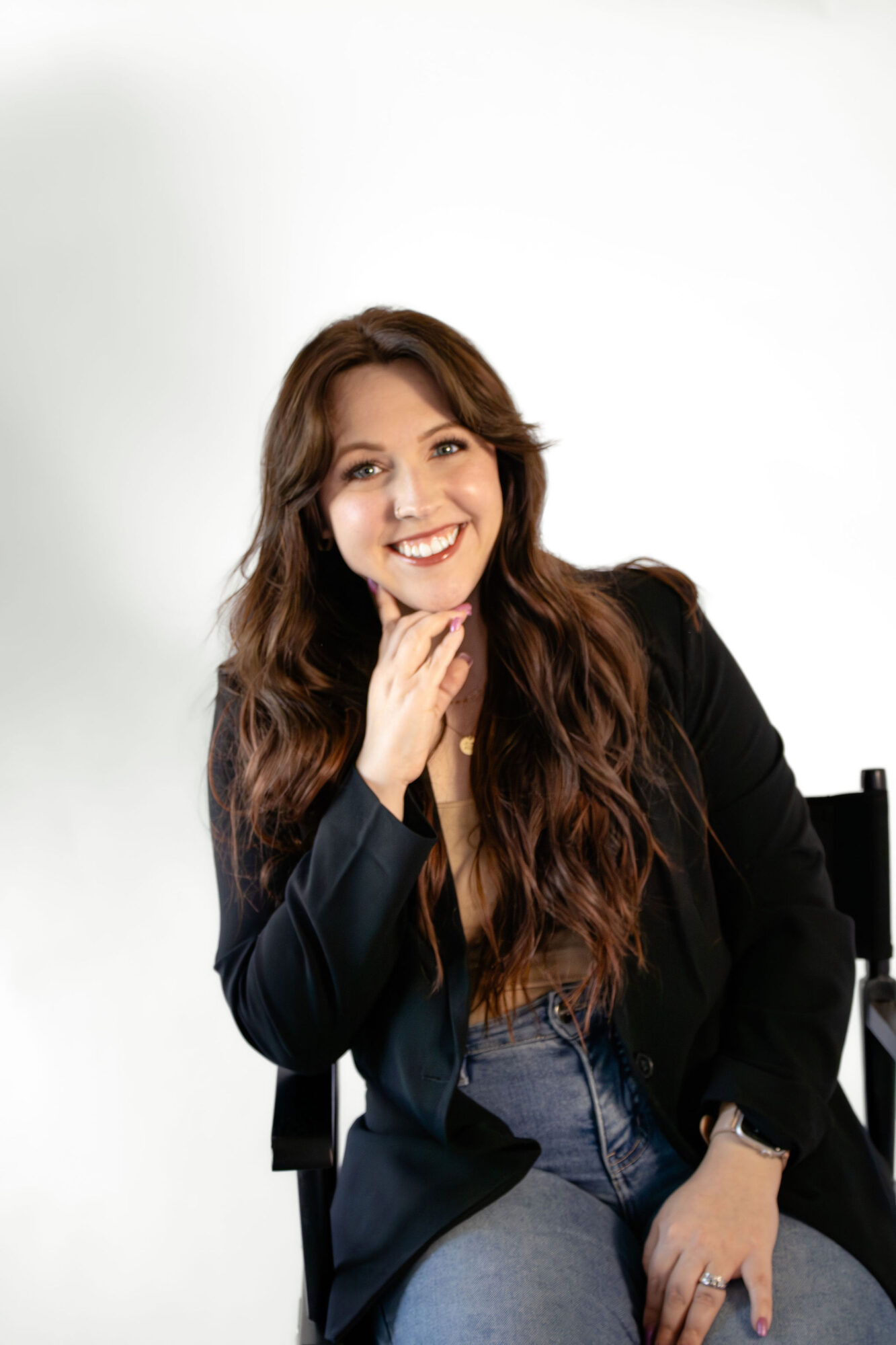 Young woman with long wavy hair smiling, wearing a black blazer and jeans, sitting against a plain white background.