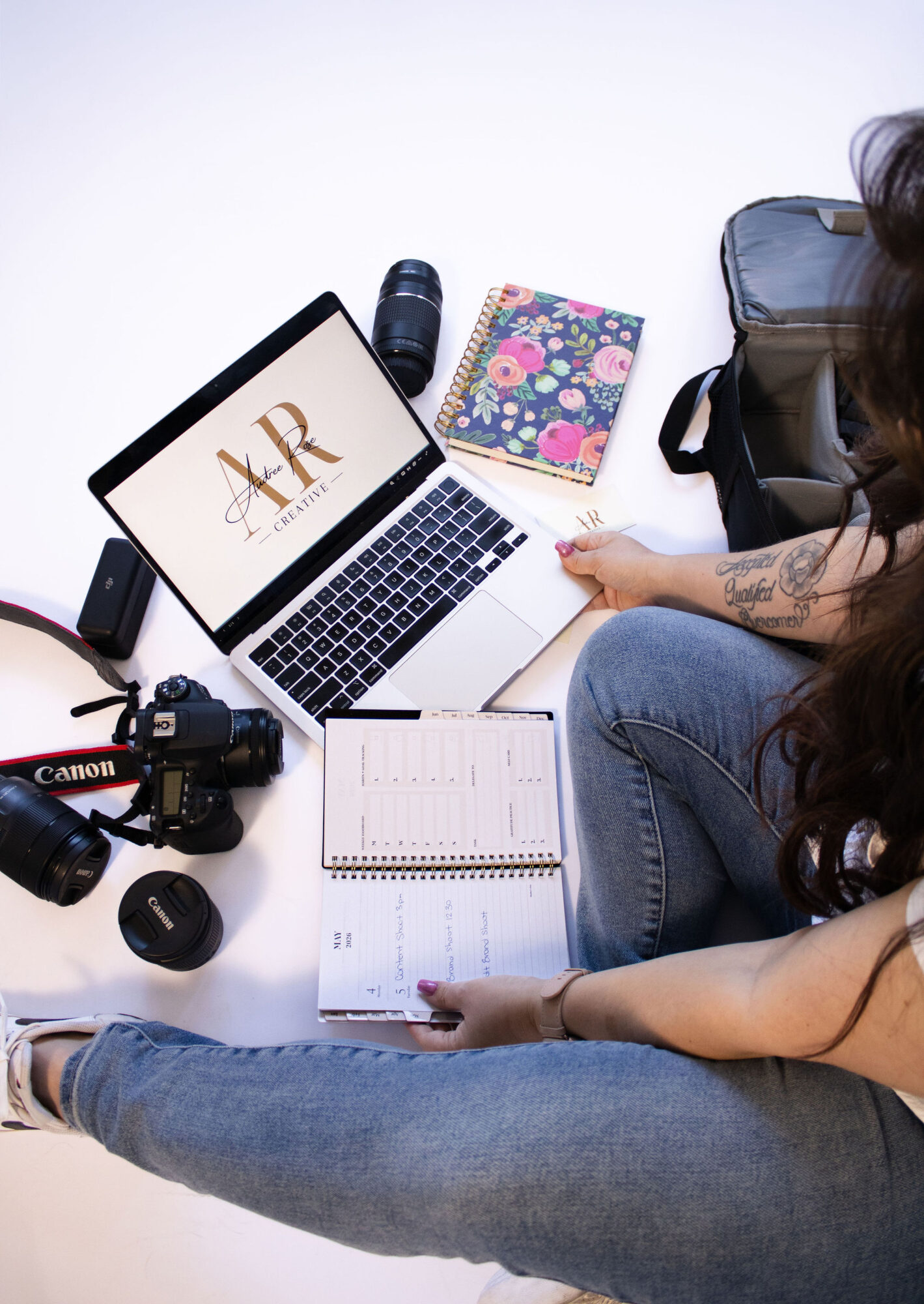 Person working at a desk with a laptop, camera, planner, and colorful notebook, viewed from above.