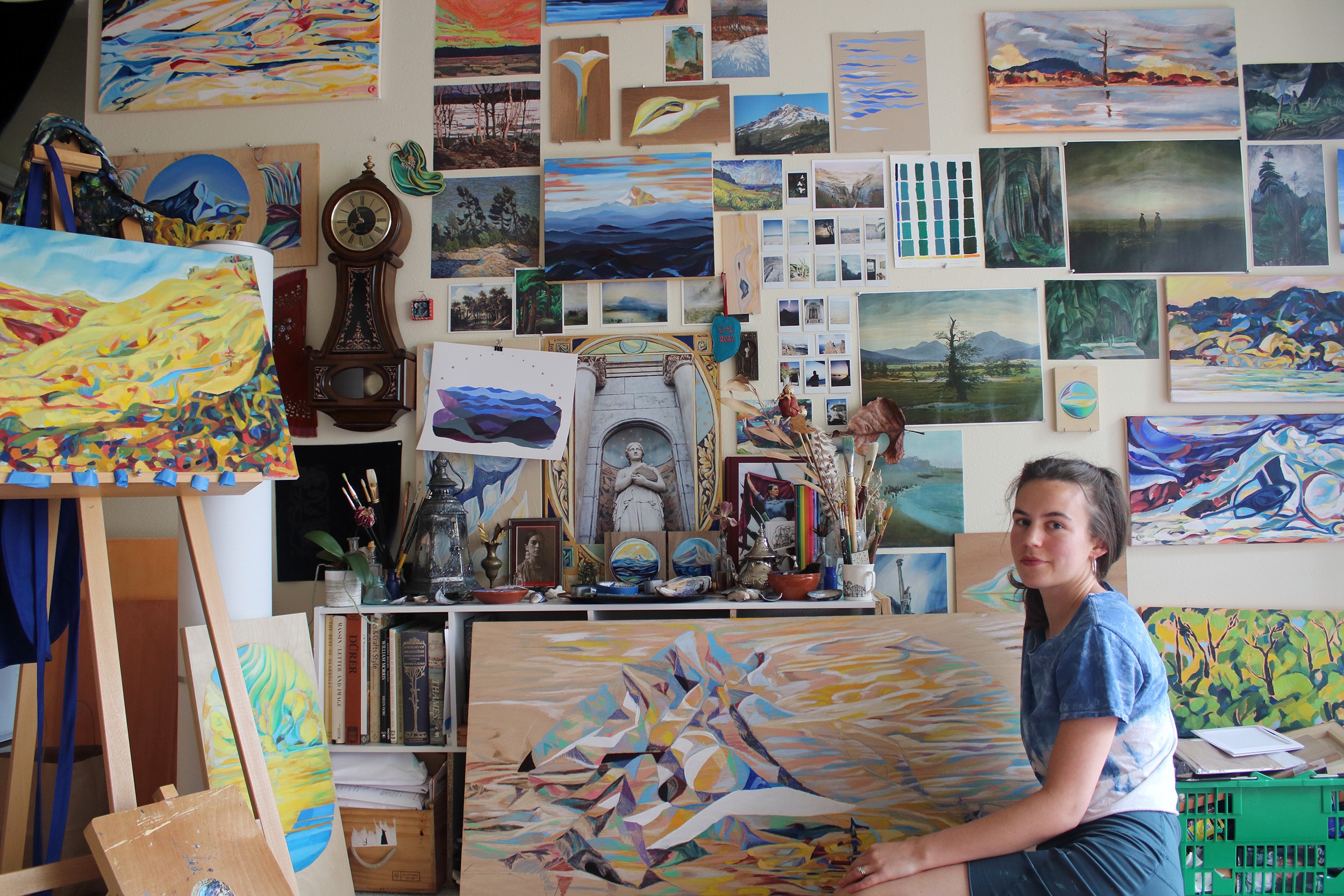 Young woman sitting in art studio surrounded by paintings and art supplies, wall covered with landscape and abstract artworks.