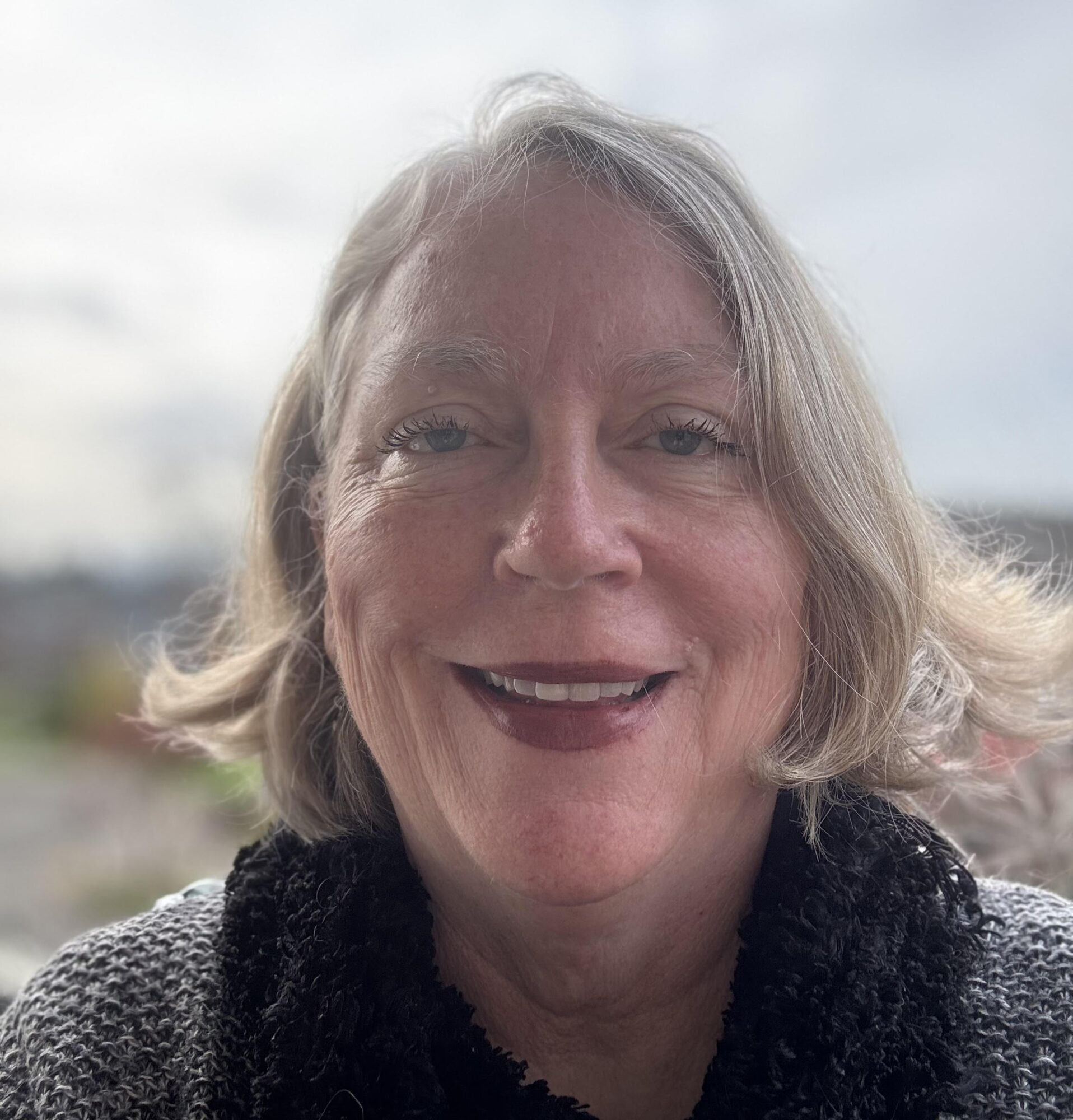 Smiling woman with shoulder-length gray hair outdoors, wearing a dark scarf and gray sweater.