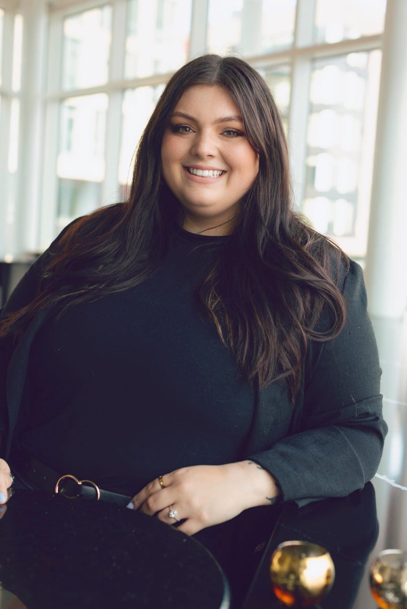 Smiling woman with long dark hair in a black top, sitting indoors near large windows, with a table and a gold object in front.