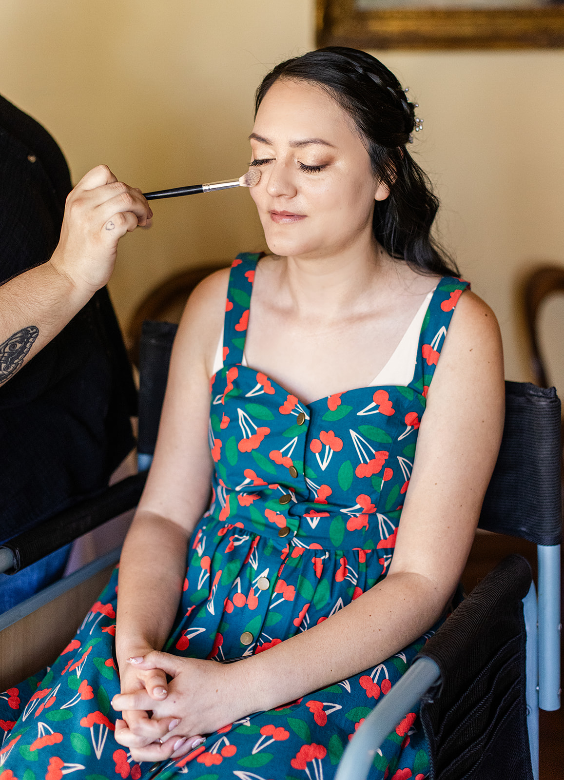Woman with closed eyes getting makeup applied, wearing a colorful dress with a pattern, seated with hands clasped.