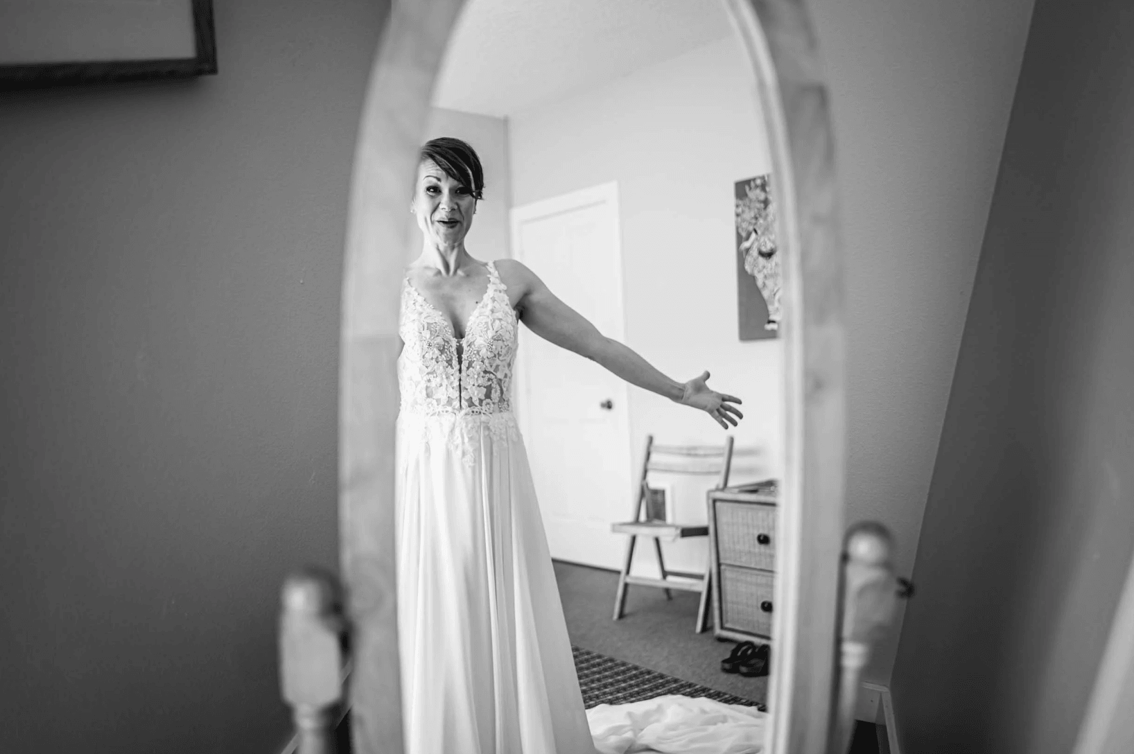Woman in a wedding dress looking at herself in a mirror, with a room and furniture visible in the background.