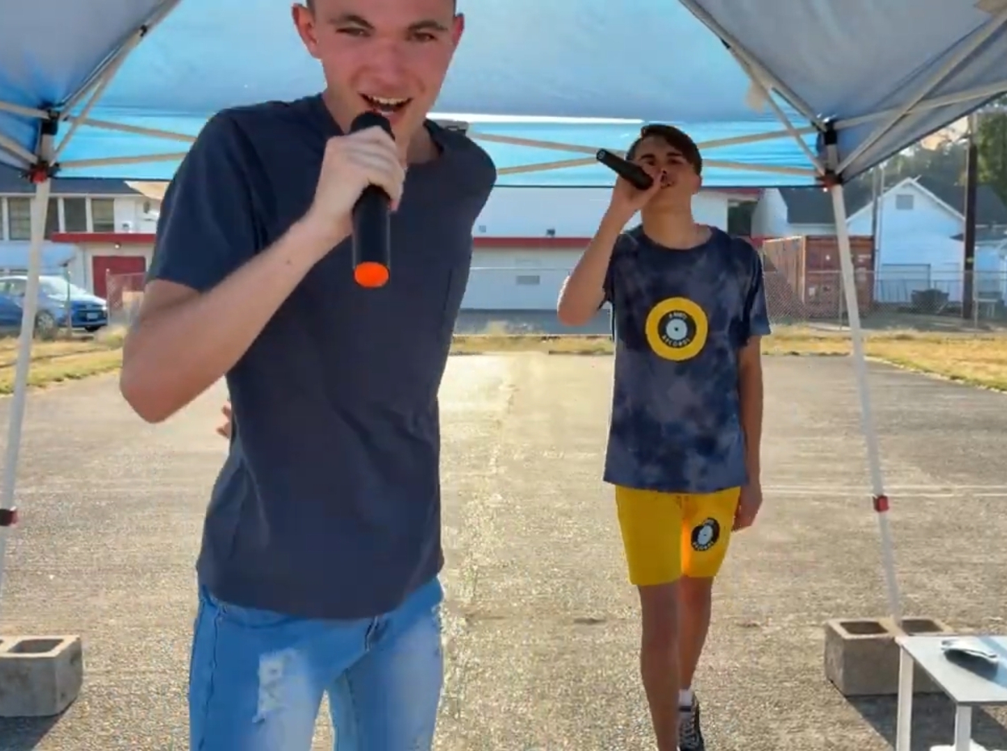 Two boys holding microphones under a canopy outdoors, one smiling at the camera, the other in the background. Bright sunny day.