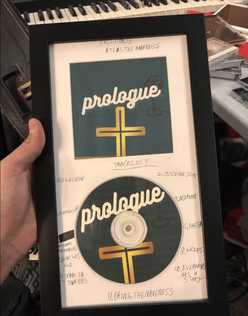 Framed CD with handwritten notes around it, held in a person's hand, background shows a keyboard and cluttered desk.