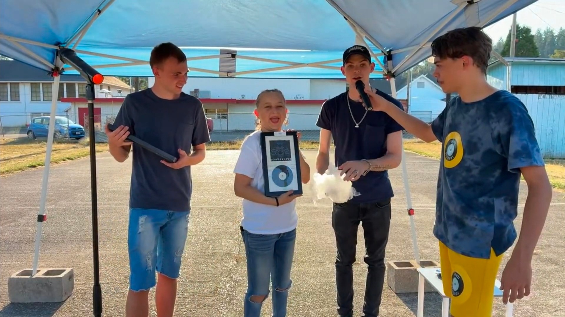 Four boys stand under a tent outdoors, one holding a microphone, with houses and trees in the background.