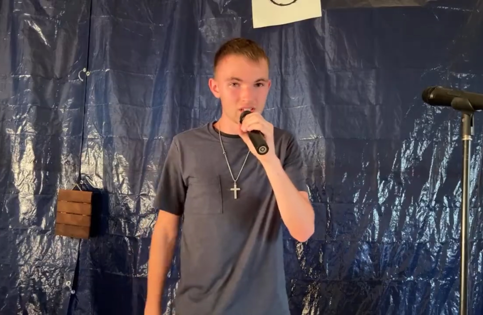 Young person holding microphone, standing in front of blue backdrop, wearing a cross necklace, speaking or singing.