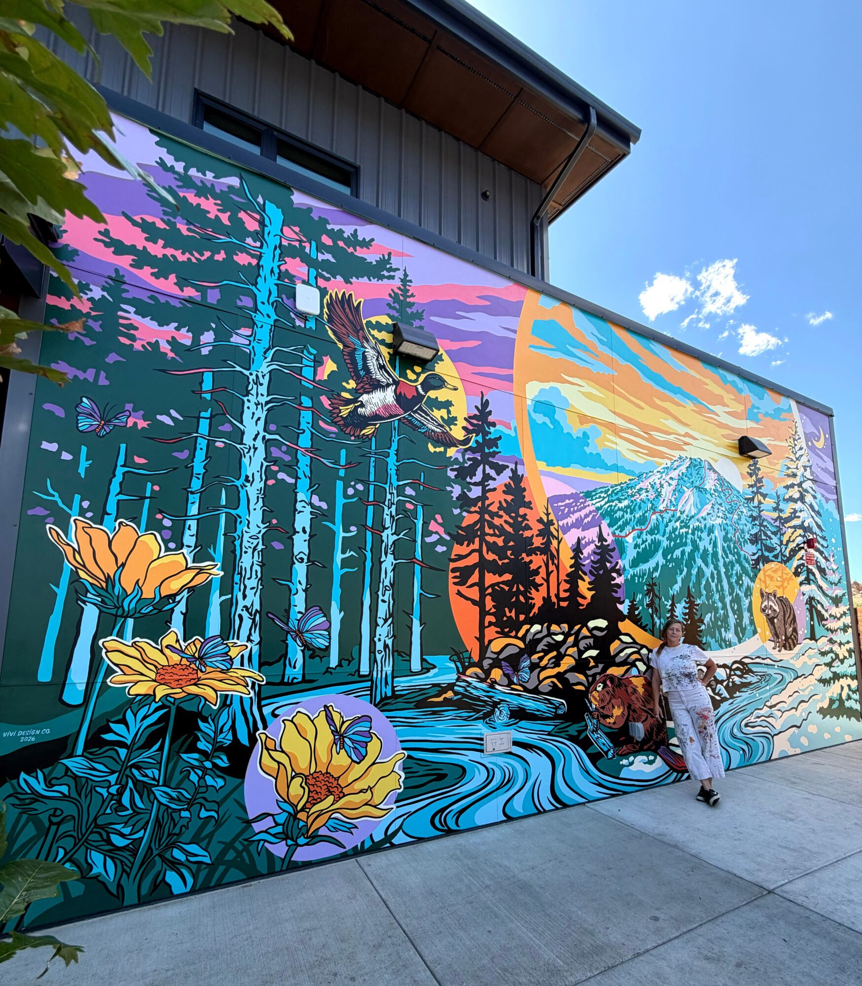 Large colorful mural depicting a forest, waterfall, mountains, and flowers with a person standing nearby.