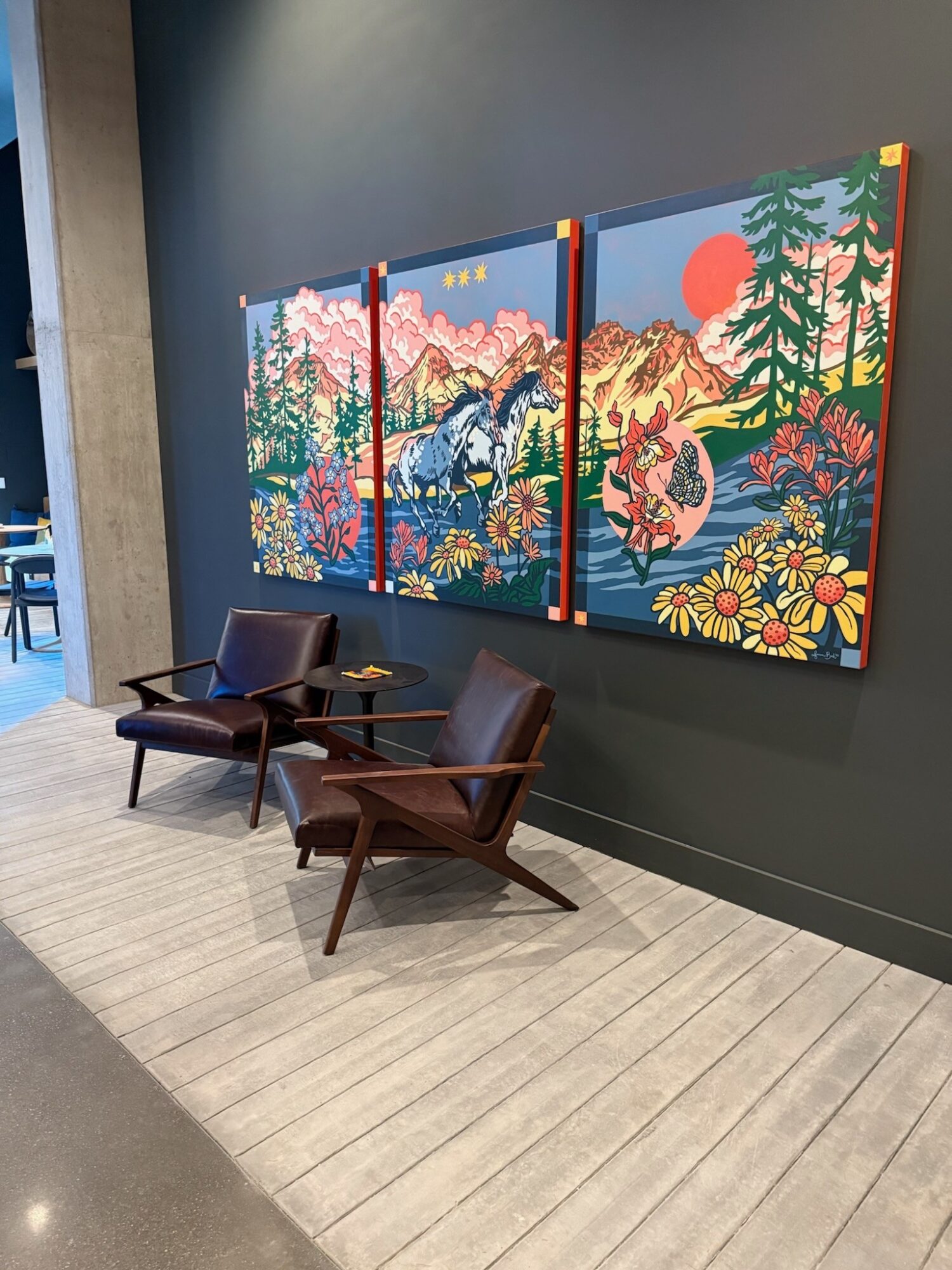 Three colorful landscape paintings on a dark wall, with two chairs and a small table in front.