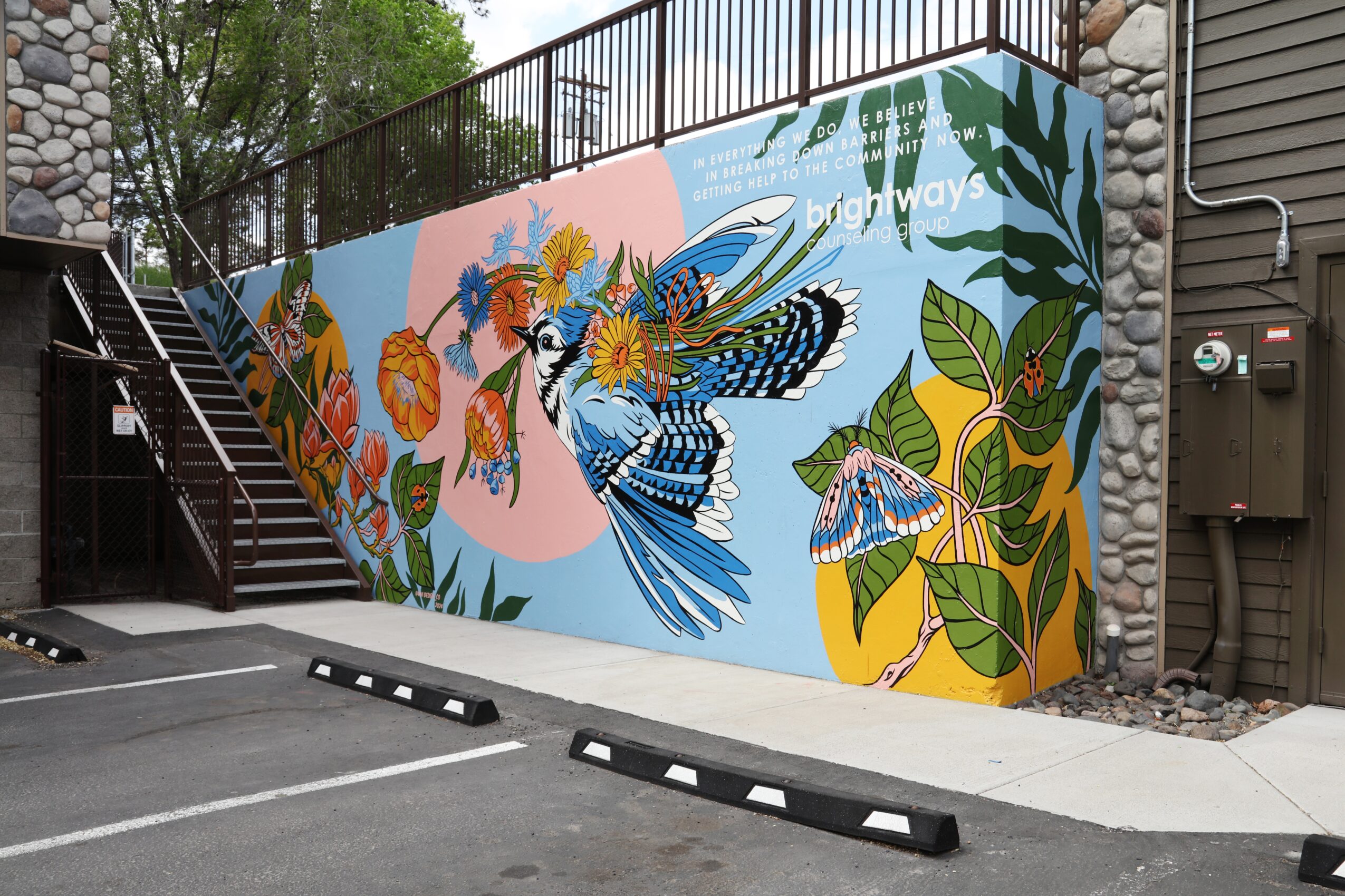 Colorful mural featuring a large blue and black bird surrounded by vibrant leaves and flowers, painted on a wall near stairs.