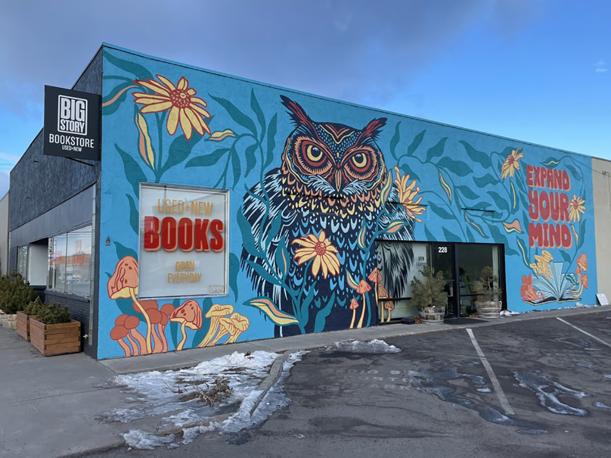 Colorful mural of an owl with flowers on a building exterior, with a bookstore sign and parking lot in front.