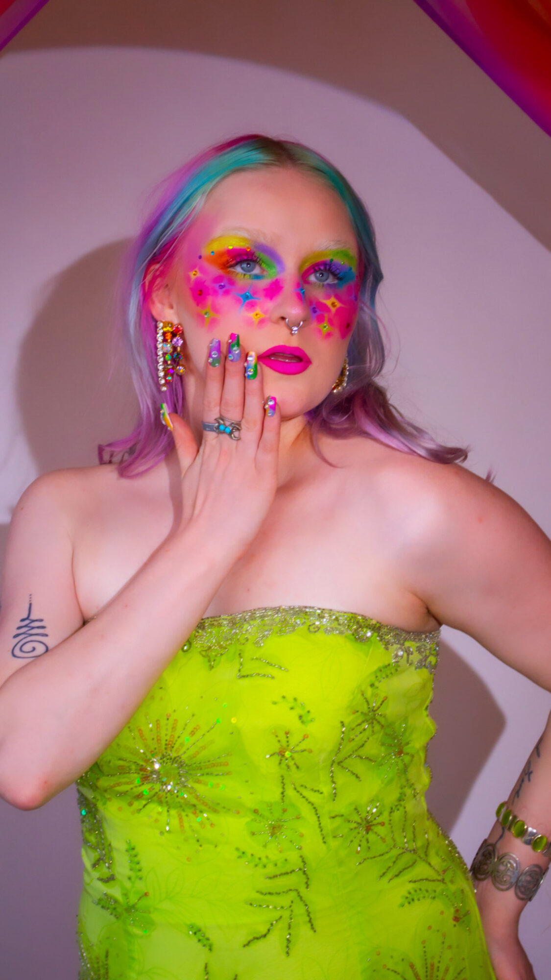 Person with colorful makeup and rainbow hair, wearing a bright yellow strapless dress, touching face with hand, against a plain background.