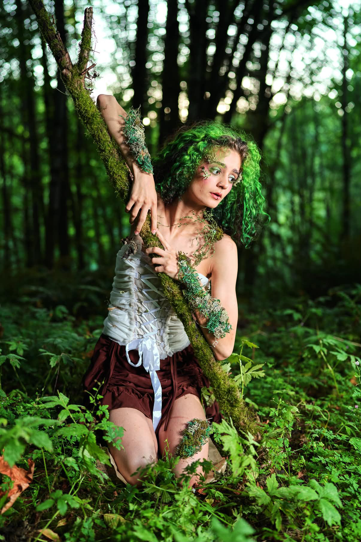 Young woman with curly green hair and tattoos kneels in a forest, holding a large stick, surrounded by green foliage.