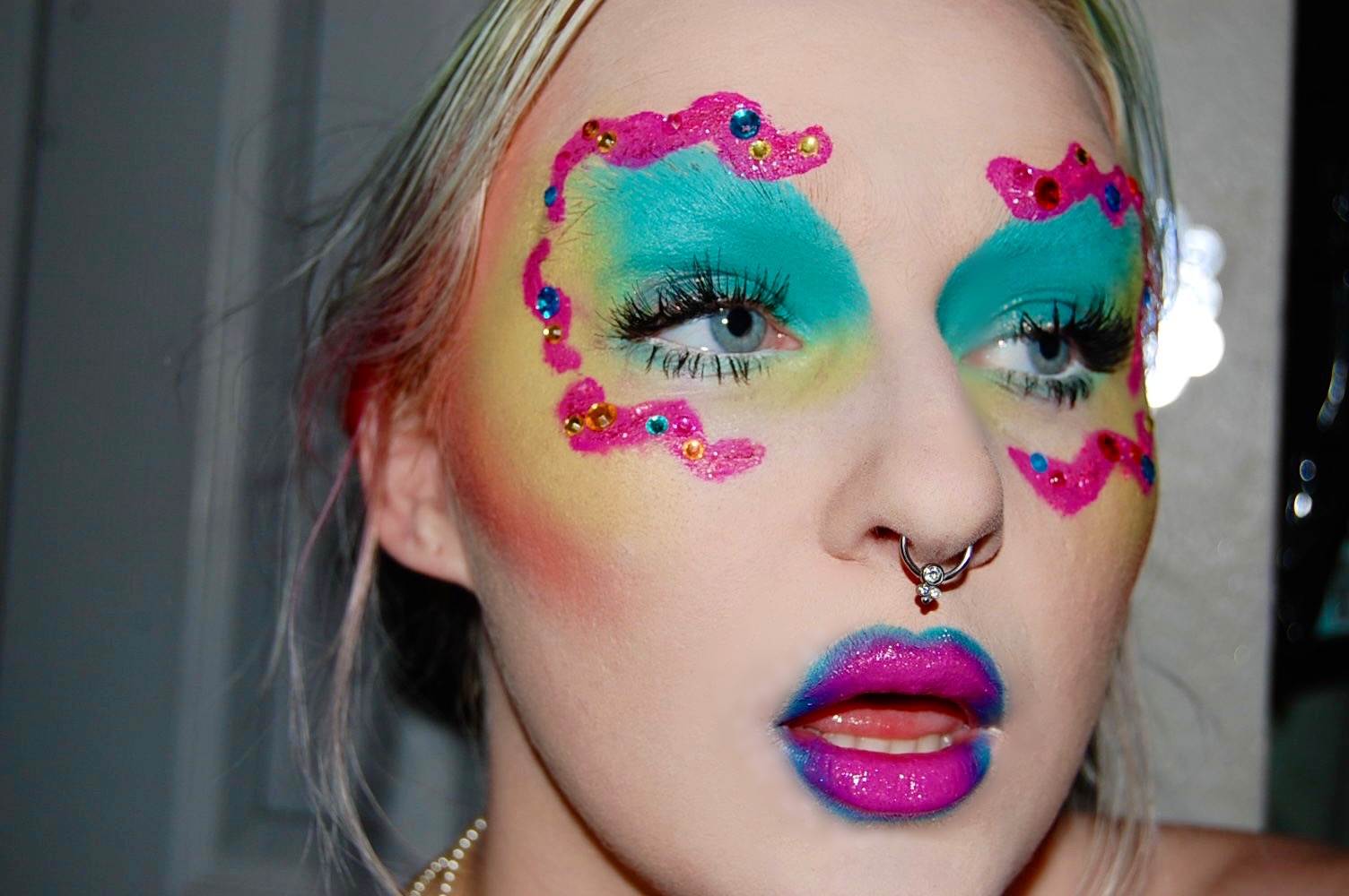 Person with colorful makeup and pink embellishments around eyes, blue lipstick, and a septum piercing.