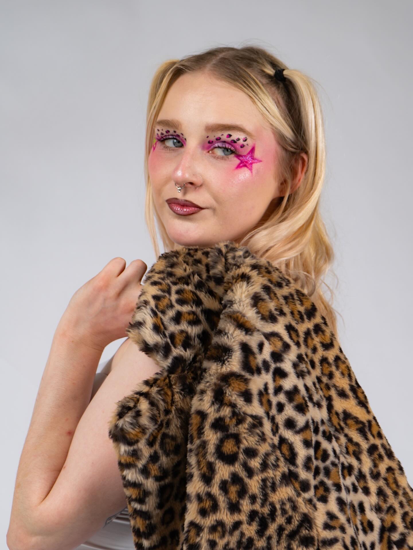 Young woman with pink star and glitter face makeup, wearing a leopard print fur coat, looking at camera.