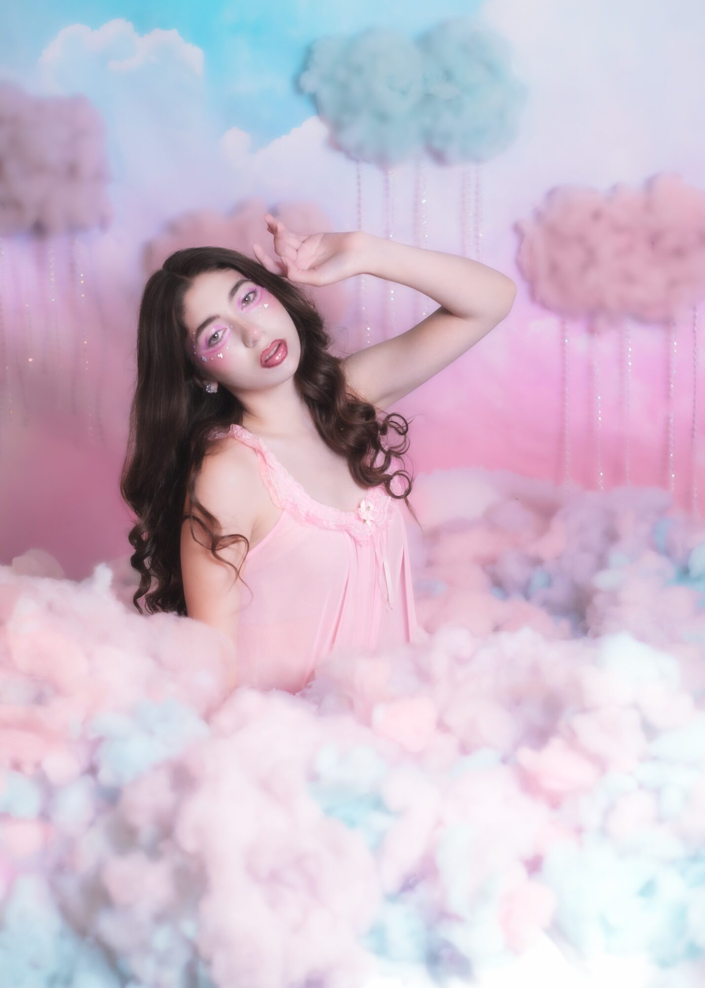 Young woman with long dark hair in pink dress surrounded by pastel-colored cotton candy clouds and hanging clouds background.