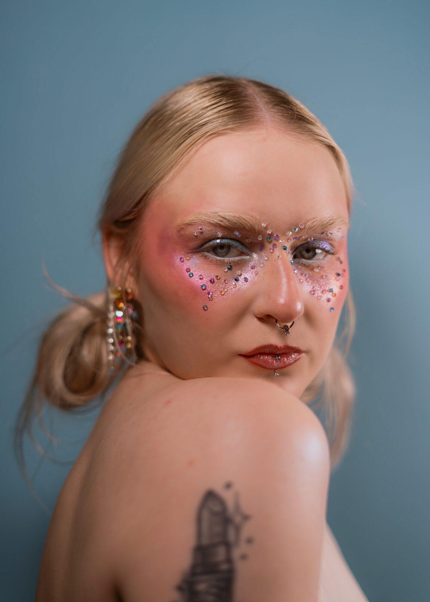 Person with glitter and face paint, looking over shoulder, with a tattoo on arm, against blue background.