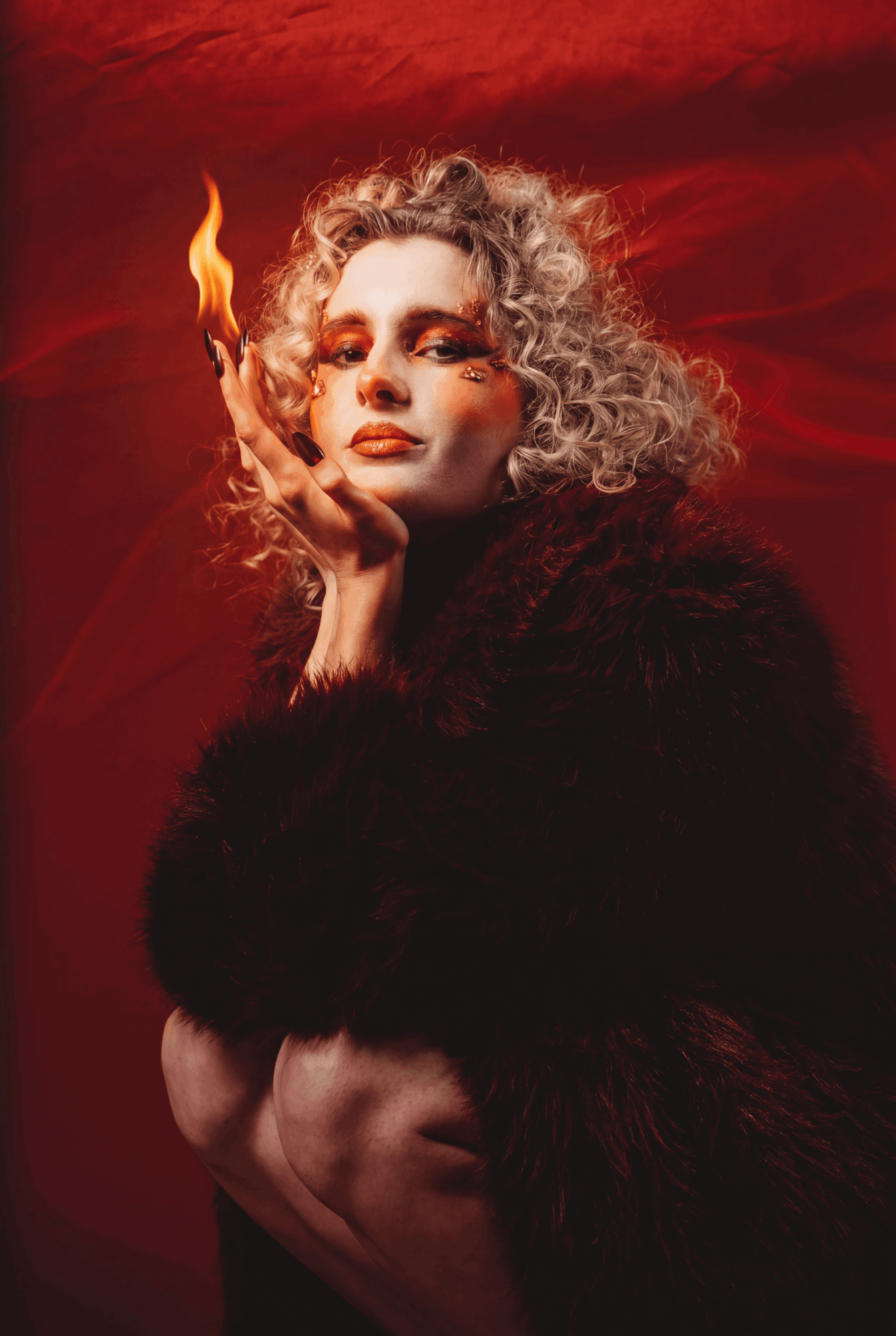 Woman with curly hair and makeup, holding a flame near her face, against a red background.