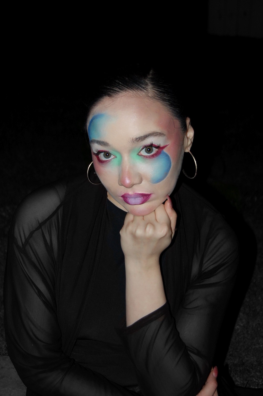 Woman with colorful makeup and hoop earrings, resting her chin on her hand, wearing a black sheer top.