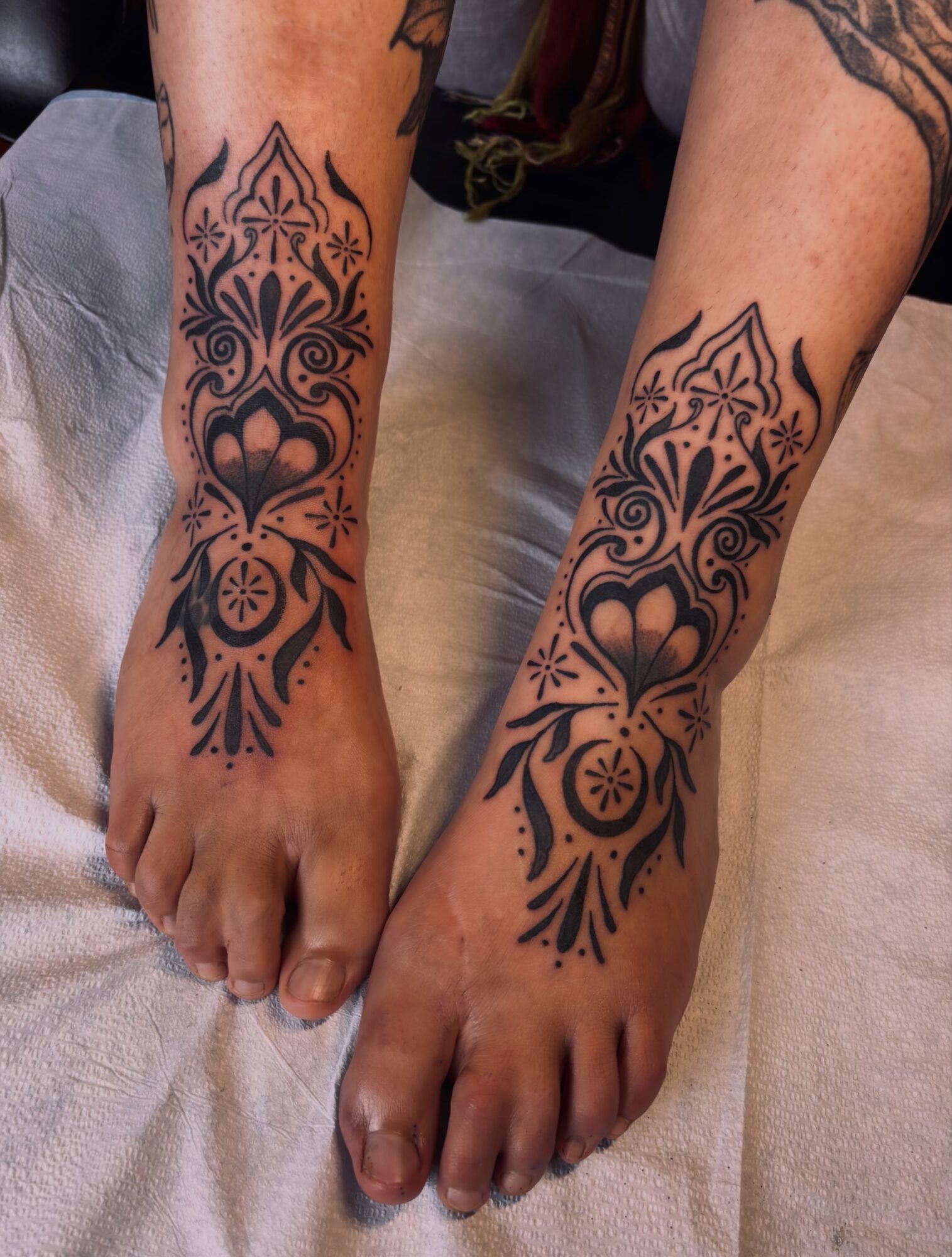 Two feet with symmetrical black tattoo designs on ankles and lower legs, featuring floral and geometric patterns.