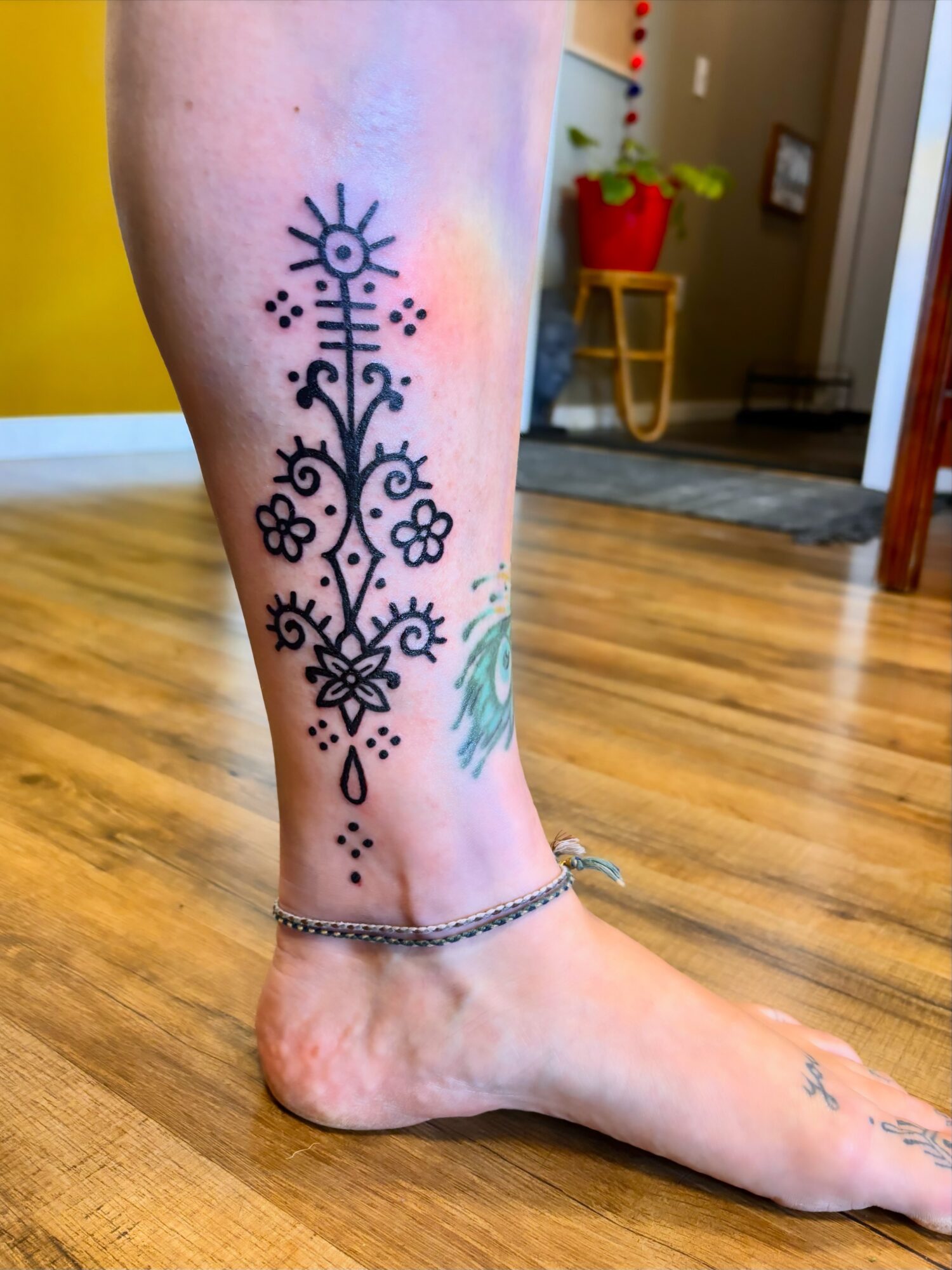 Tattoo on ankle with floral and geometric patterns, and an anklet, in a room with wooden floor and furniture.